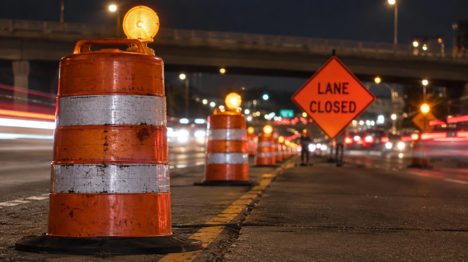 Orange construction barrels and lane closure signs along a highway at night.