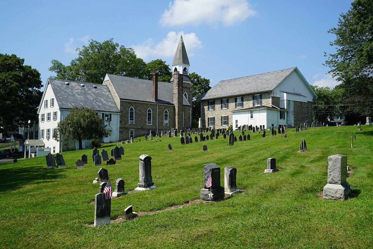 Historic cemetery with old headstones and American flags beside colonial stone church buildings.