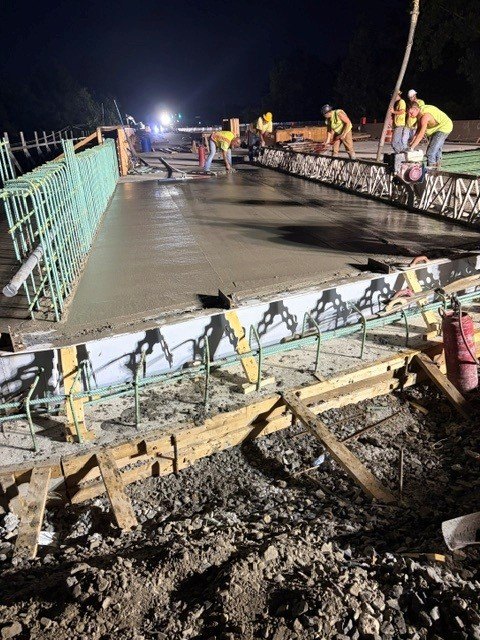 Workers in high-visibility vests smooth wet concrete on a bridge deck at night, green rebar visible along edges.