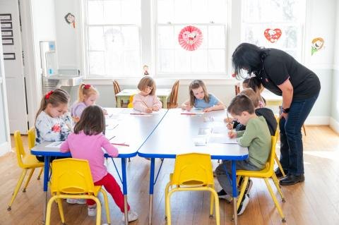 Young children draw at a classroom table while an adult instructor assists them.