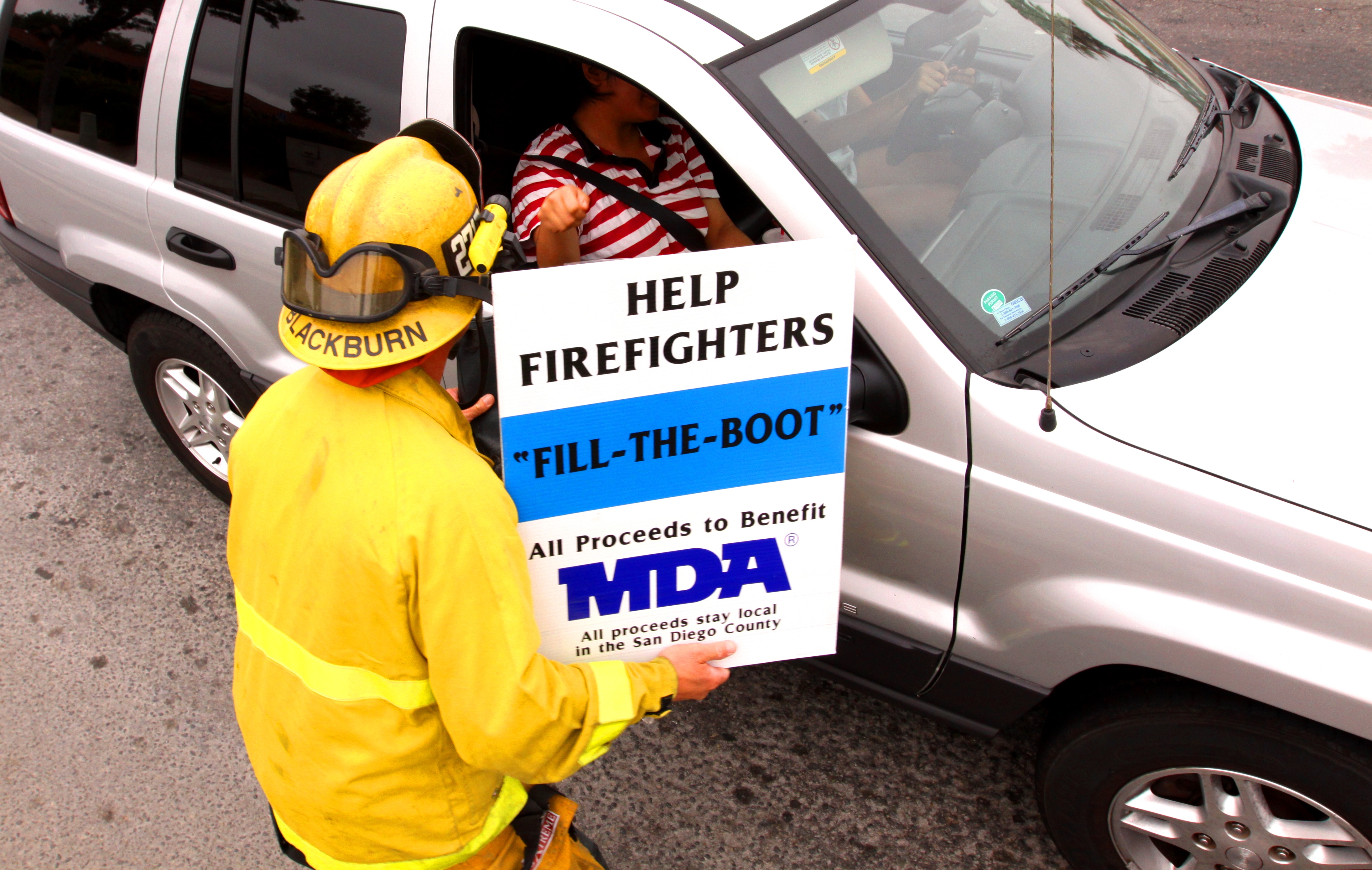 Firefighter in yellow gear holds a Fill the Boot MDA sign at a car window.