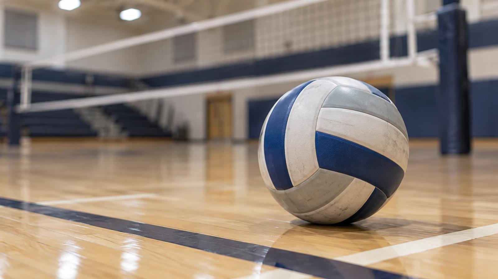 A volleyball resting on a hardwood gym floor near the net with court lines visible.
