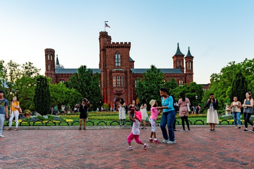 Red-brick Victorian castle building at dusk with visitors gathered on a brick plaza in front.