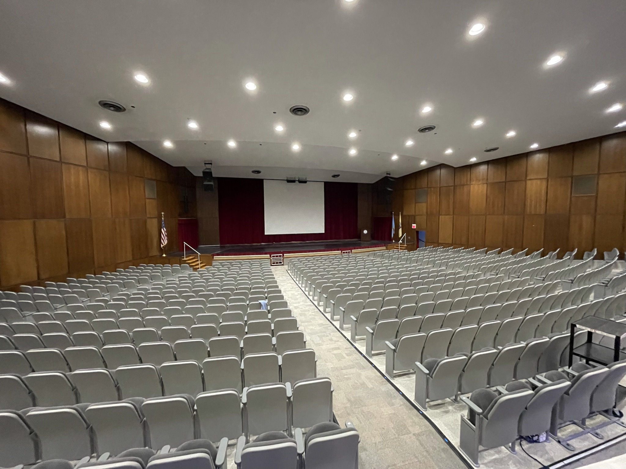 Empty school auditorium with rows of gray seats facing a stage with red curtains.