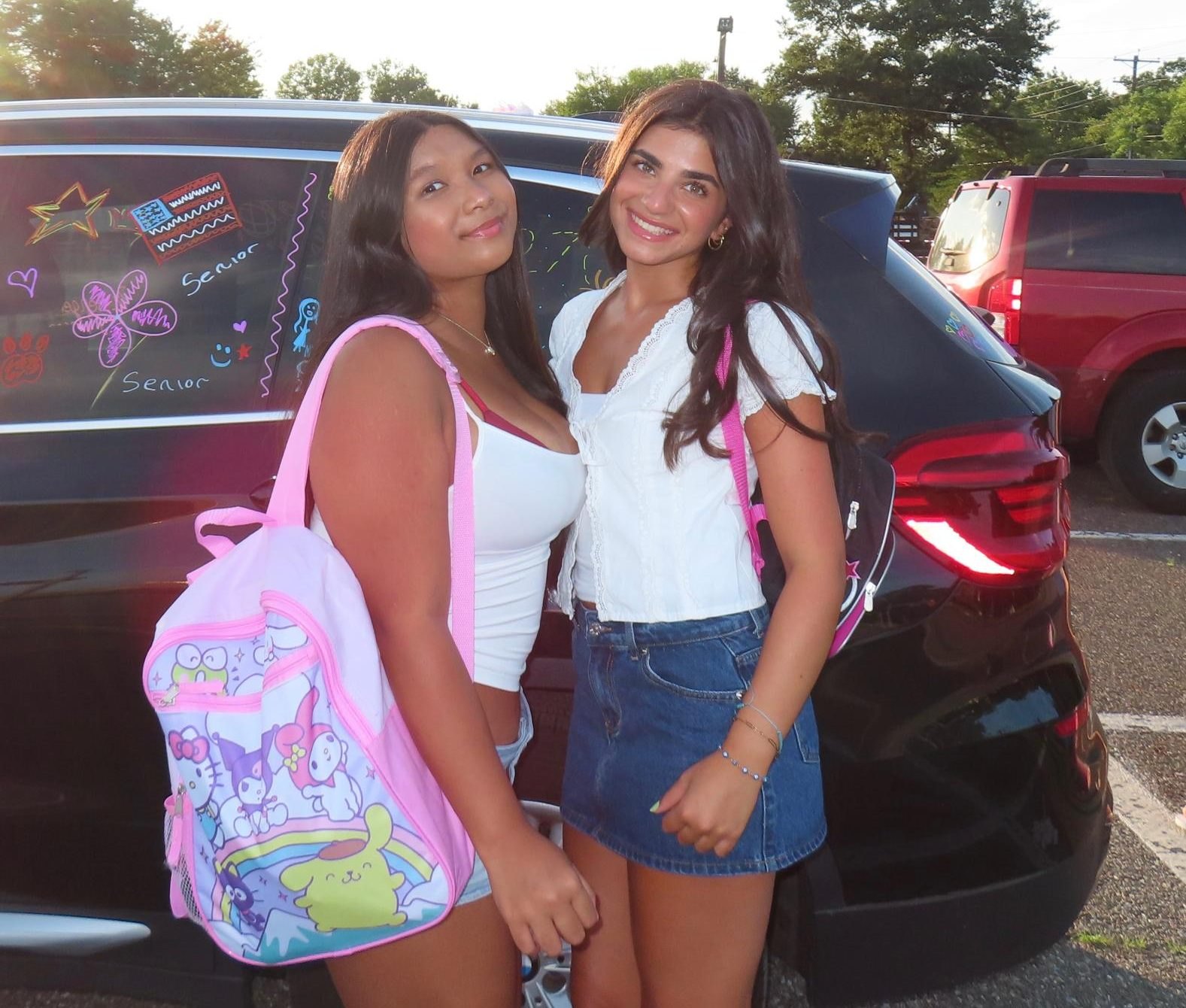 Two young women in a parking lot, one wearing a pink Sanrio character backpack, beside a car with 'Senior' written on the window.