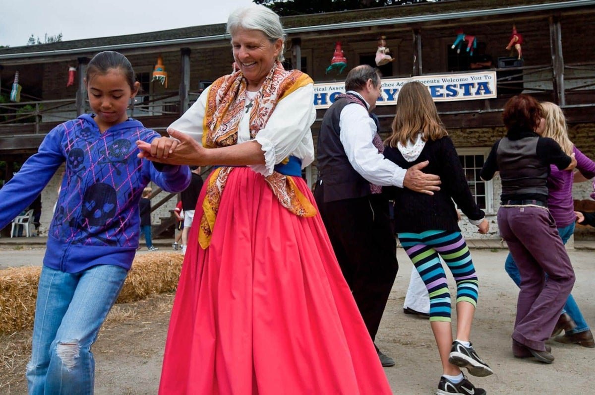 Woman in colorful traditional dress dances with a young girl outdoors at a fiesta event.