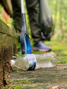 Close-up of a plastic bottle on a trail as a person with trash grabbers reaches to pick it up.