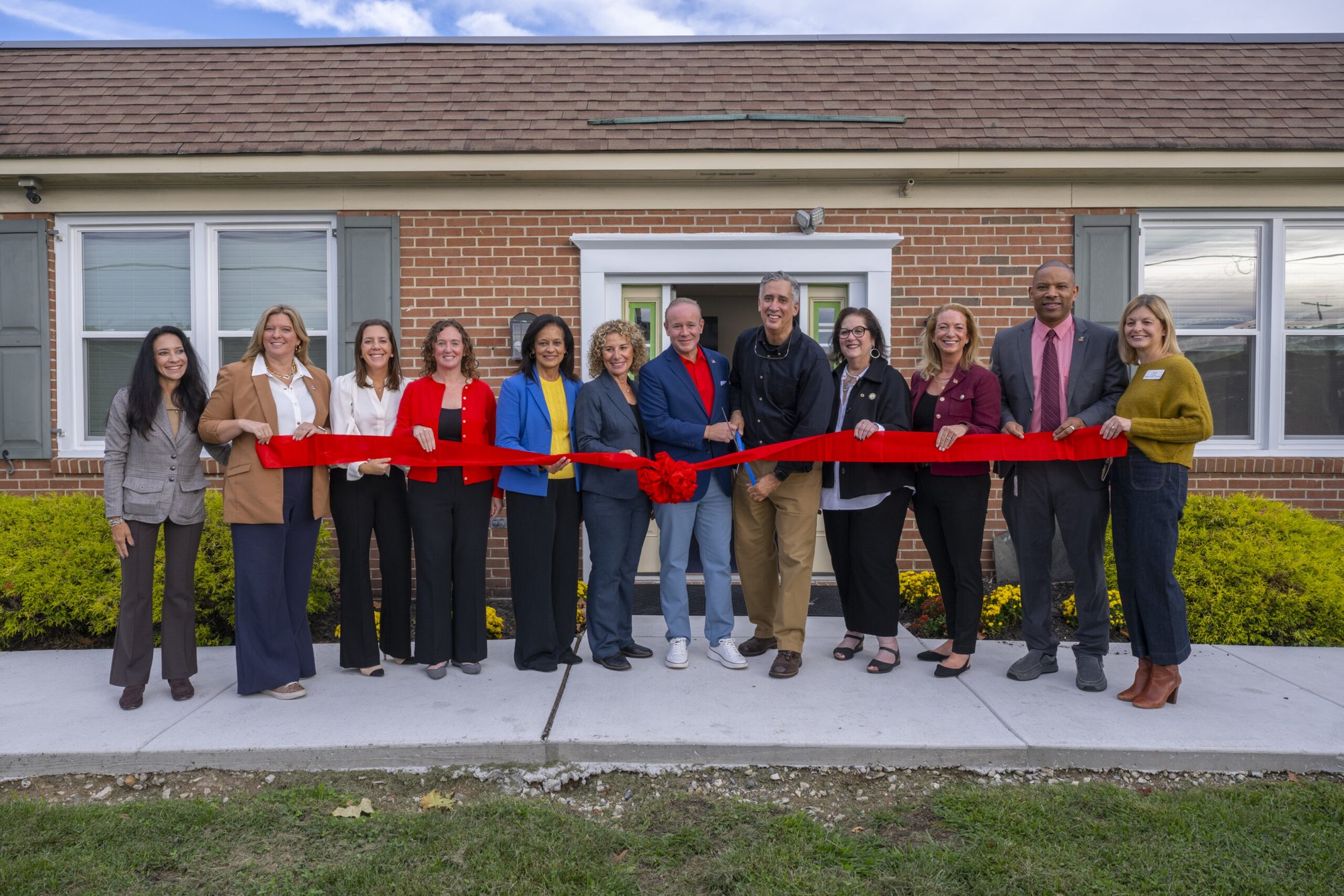 Group of adults holding a red ribbon in front of a single-story brick building.