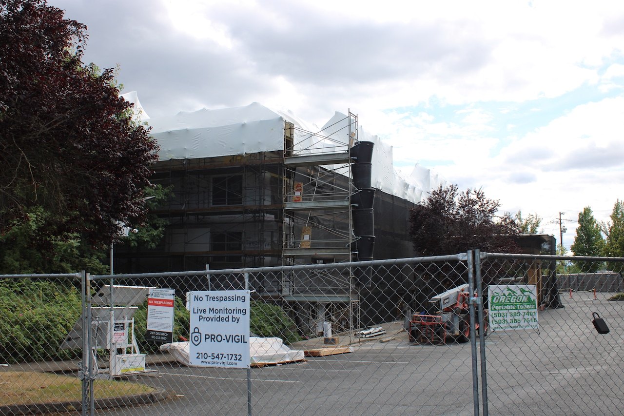 A multi-story building under renovation with scaffolding and protective sheeting, behind a chain-link security fence.