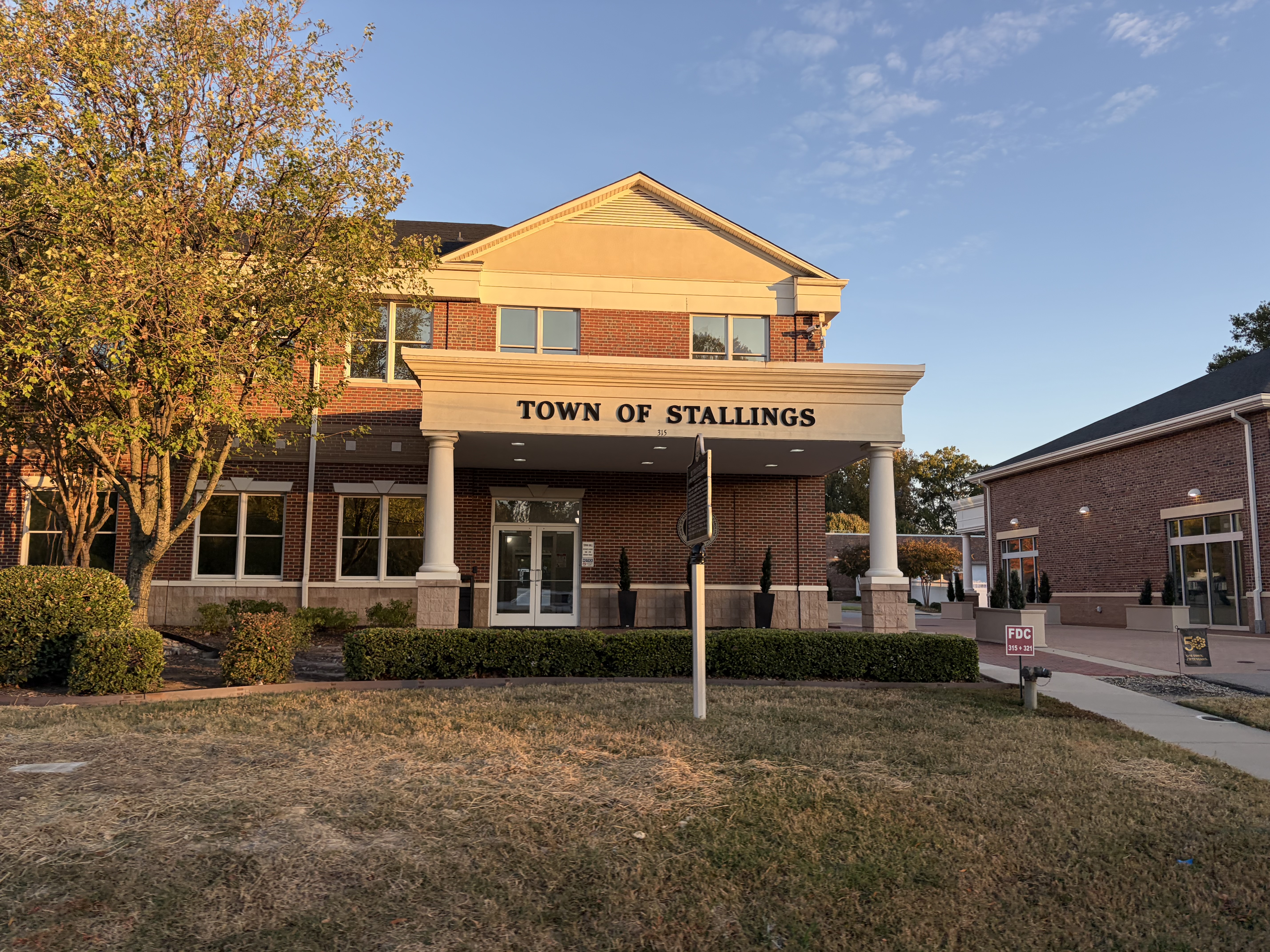 Two-story brick town hall with white columns and a flagpole on a grassy lawn.
