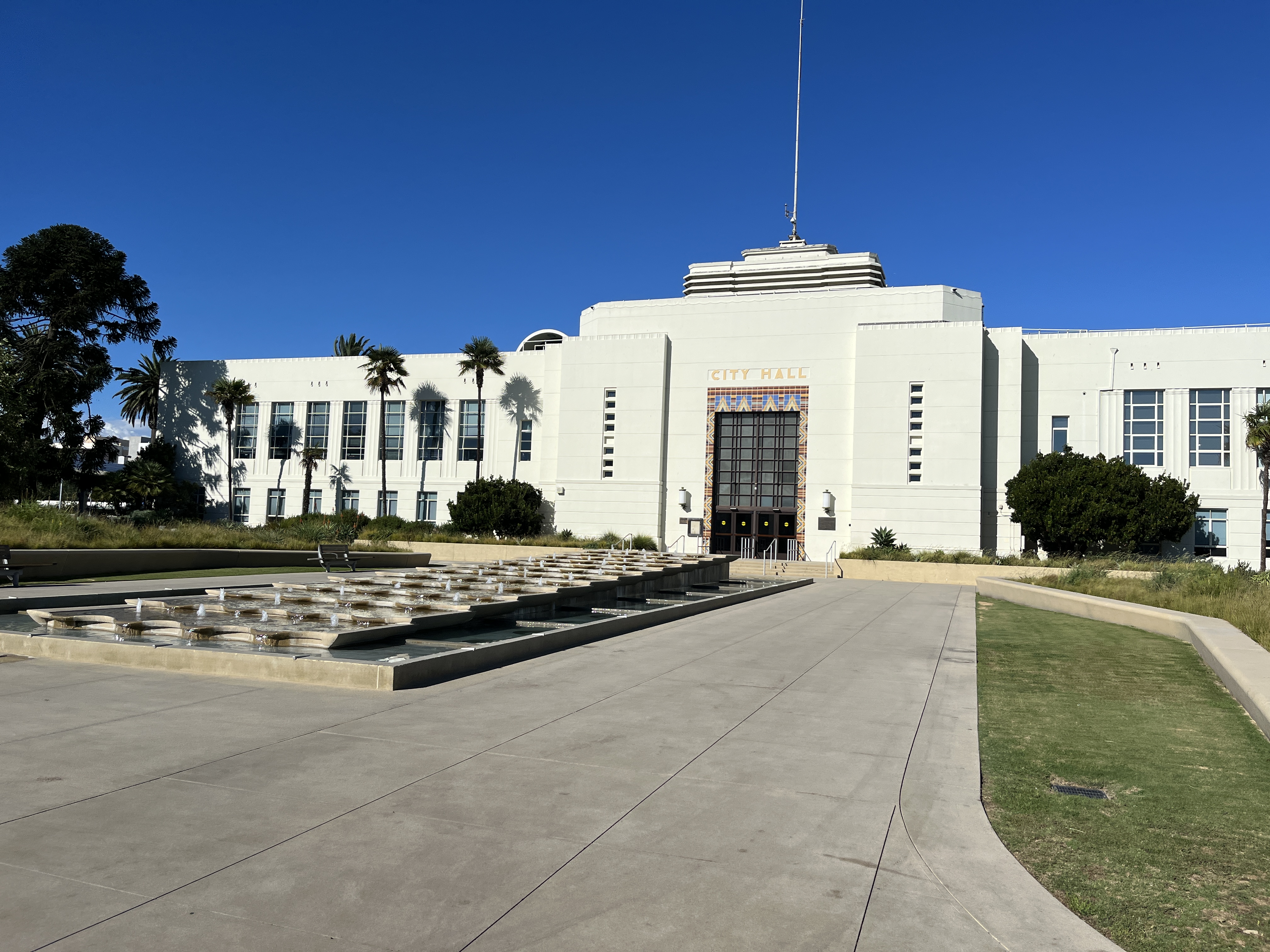 White Art Deco city hall building with palm trees and a fountain plaza in front.