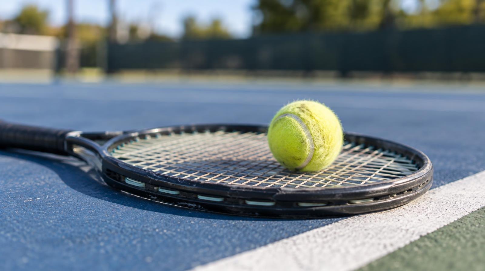 A yellow tennis ball resting on the strings of a tennis racket on a hard court.
