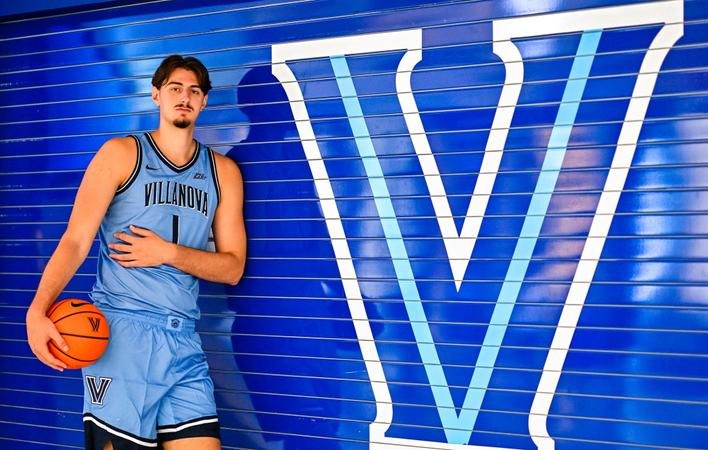 Tall basketball player in light blue Villanova uniform holding a basketball in front of a blue V logo.