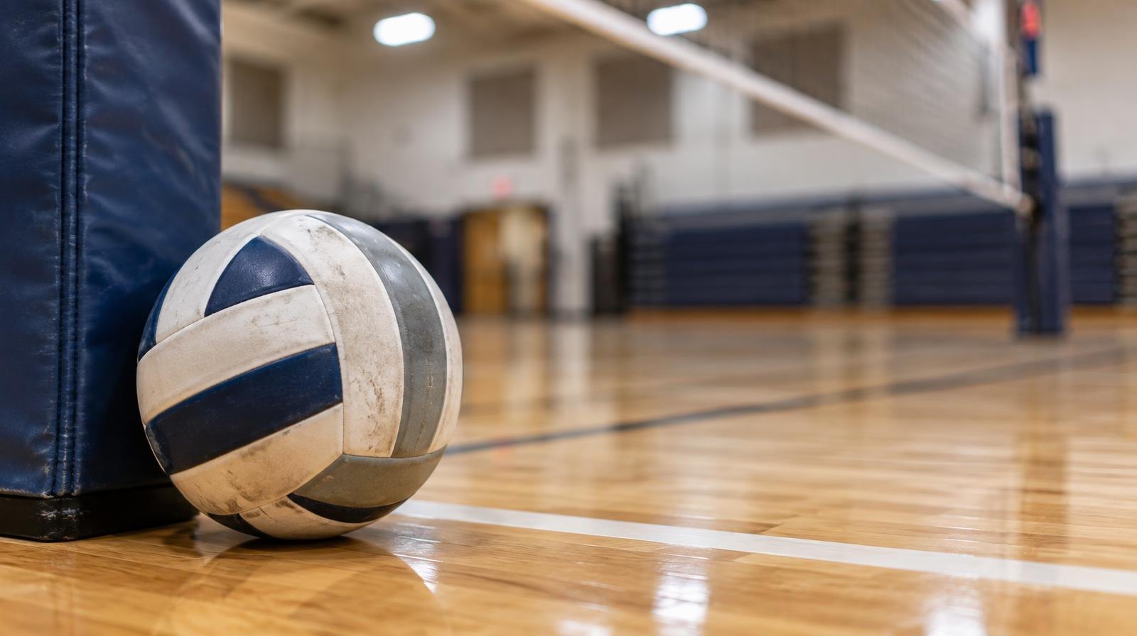 A volleyball resting on a hardwood gym floor beside a net post.