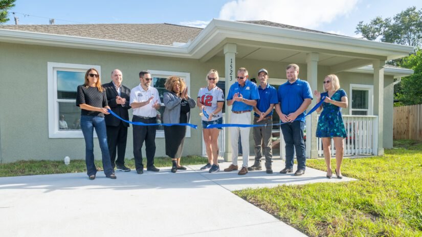 Group of people cutting a blue ribbon in front of a newly rebuilt single-family home.