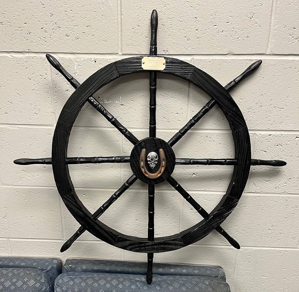 Large black wooden ship's wheel with a skull ornament at the center hub, leaning against a wall.