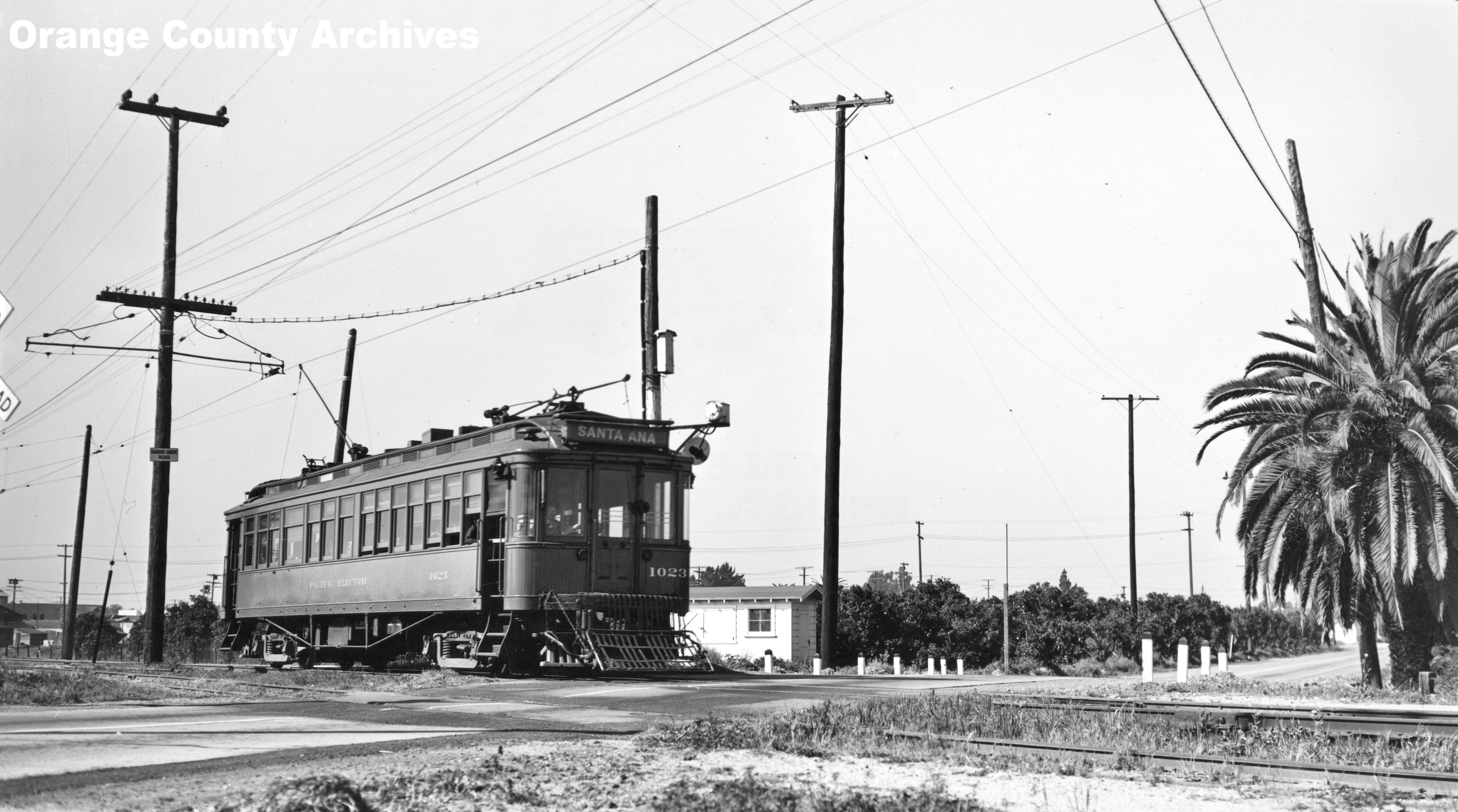 Black-and-white photo of a Pacific Electric trolley car crossing a road with utility poles and palm trees visible.