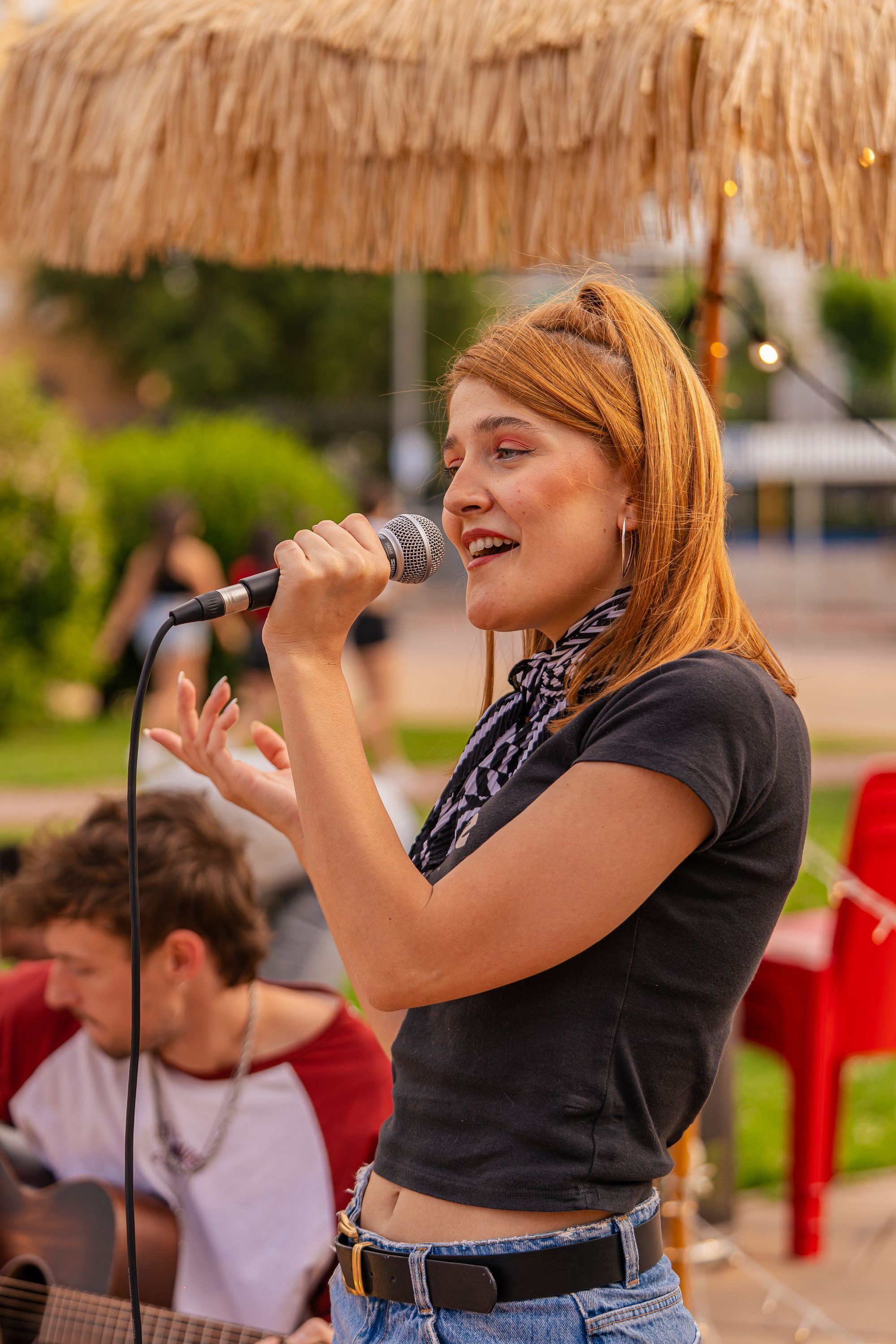 A woman sings into a microphone outdoors, with a guitarist behind her.