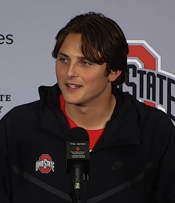 Young man in an Ohio State jacket speaking at a press conference podium.