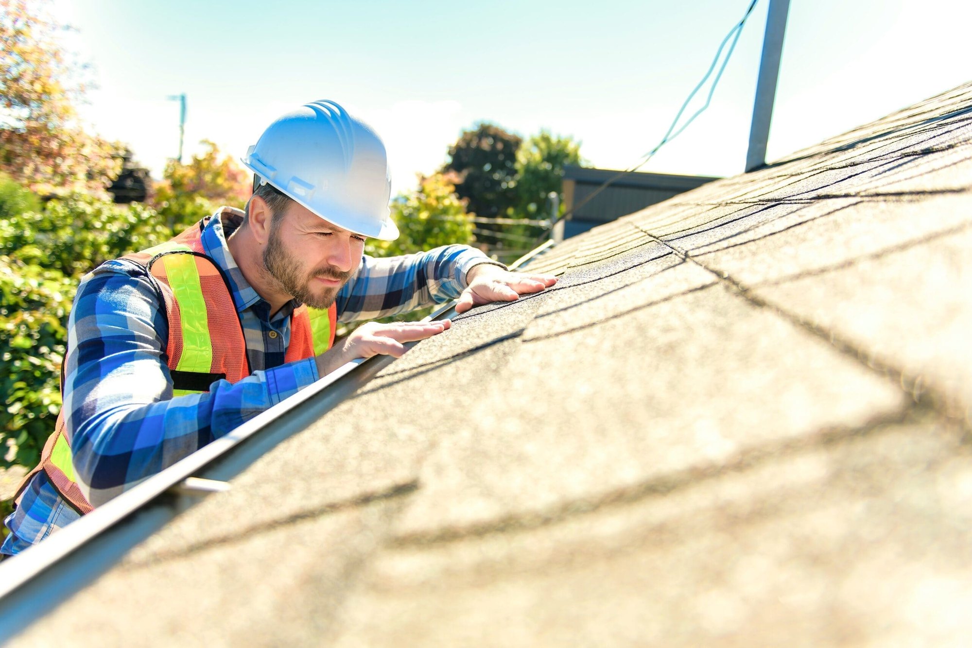 Worker in blue hard hat and orange safety vest crouching on a sloped residential roof.