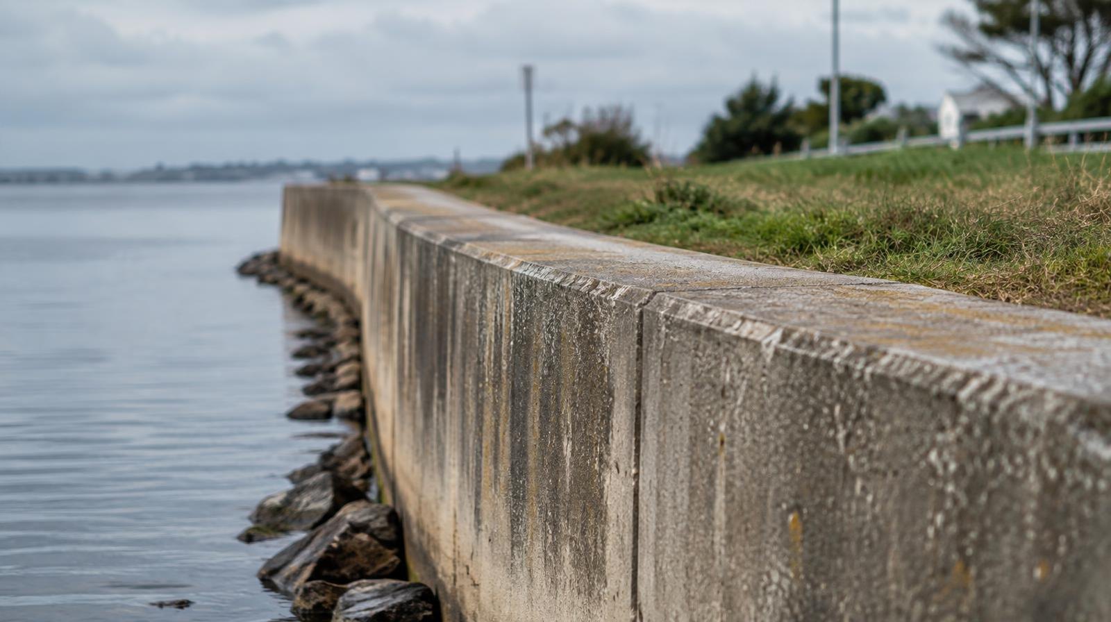 A concrete seawall runs along a calm waterfront under an overcast sky.