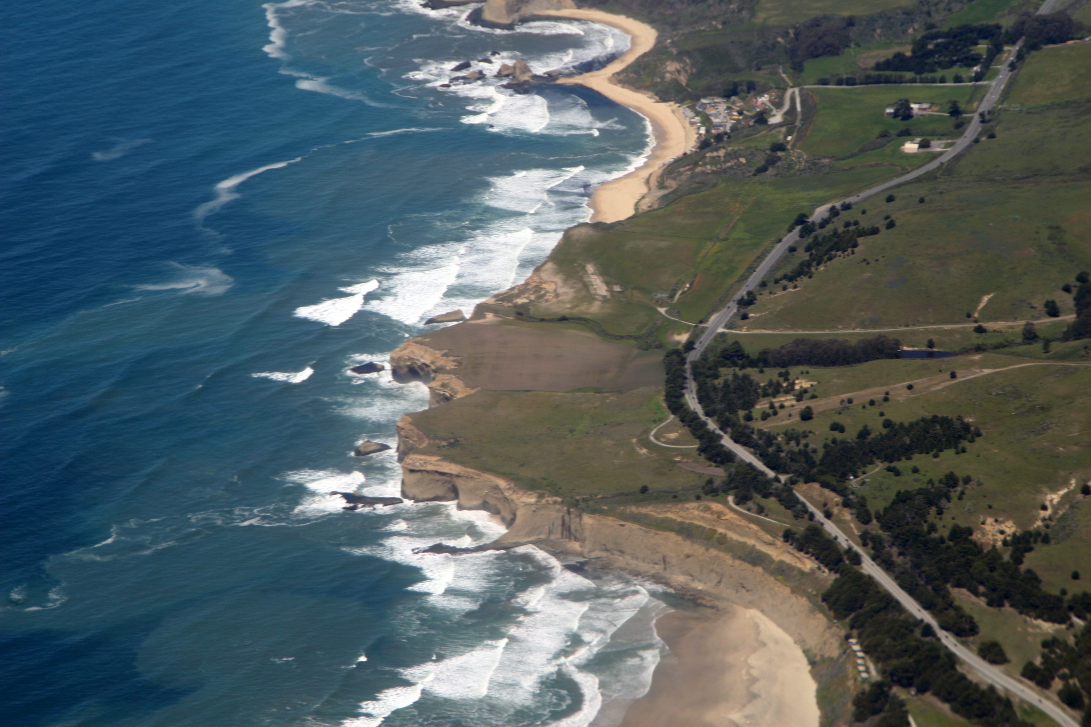 Aerial view of a rugged Pacific coastline with sandy beaches, breaking waves, and a highway along the bluffs.