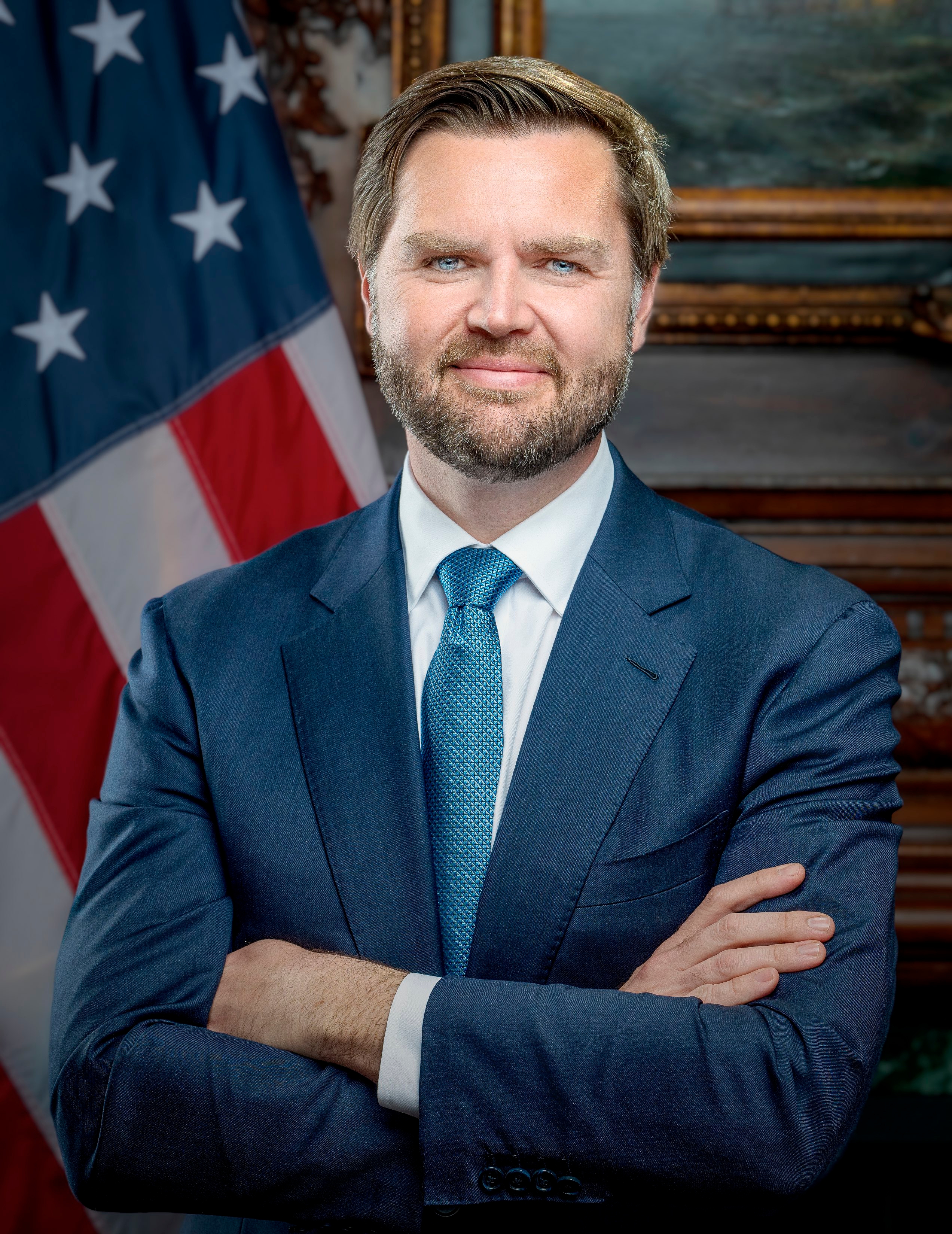 Man in navy suit with teal tie, arms crossed, standing before an American flag.