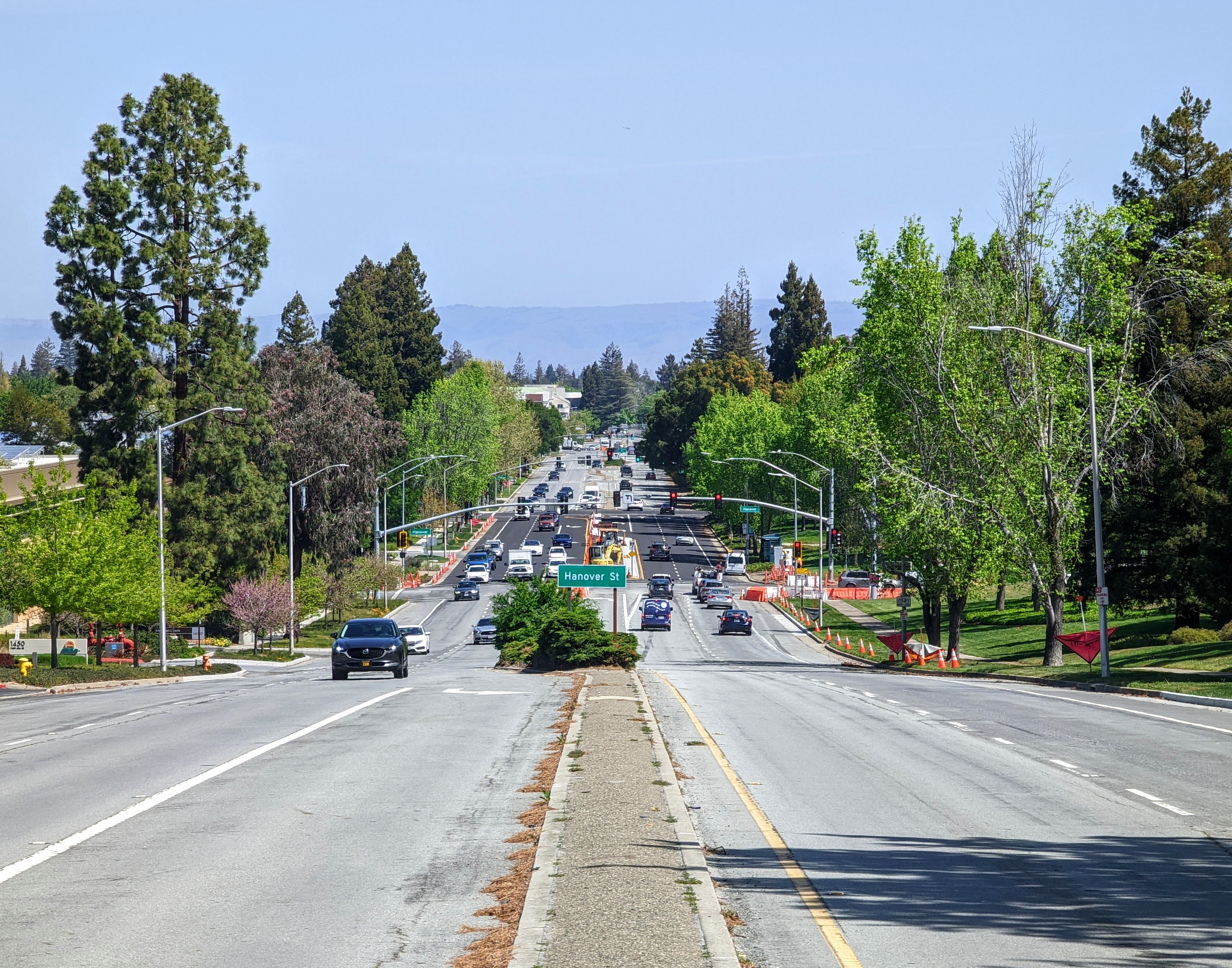 A road descending toward a busy street intersection lined with trees and utility poles.