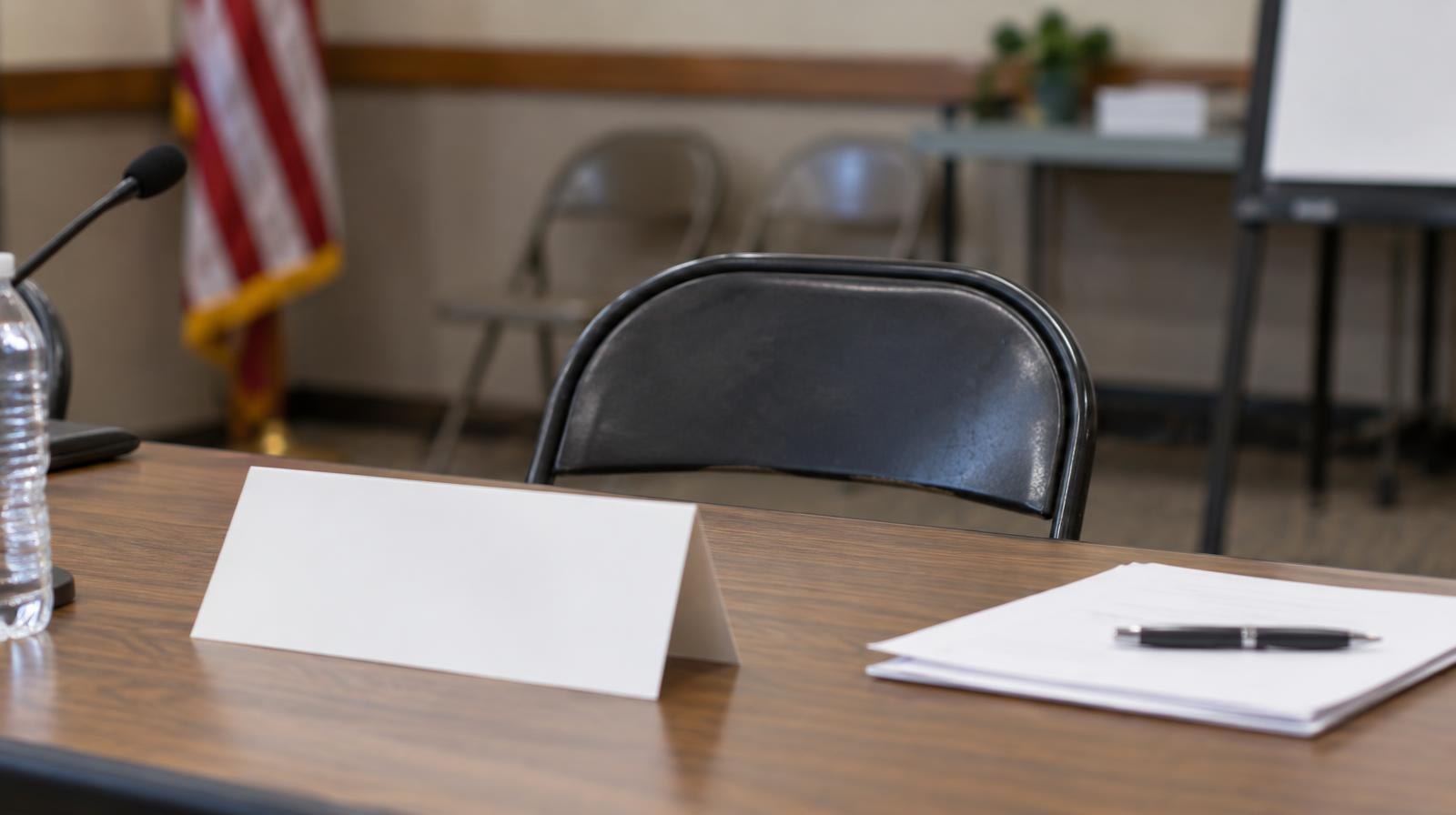 An empty folding chair at a meeting table with a blank name placard in front of it.
