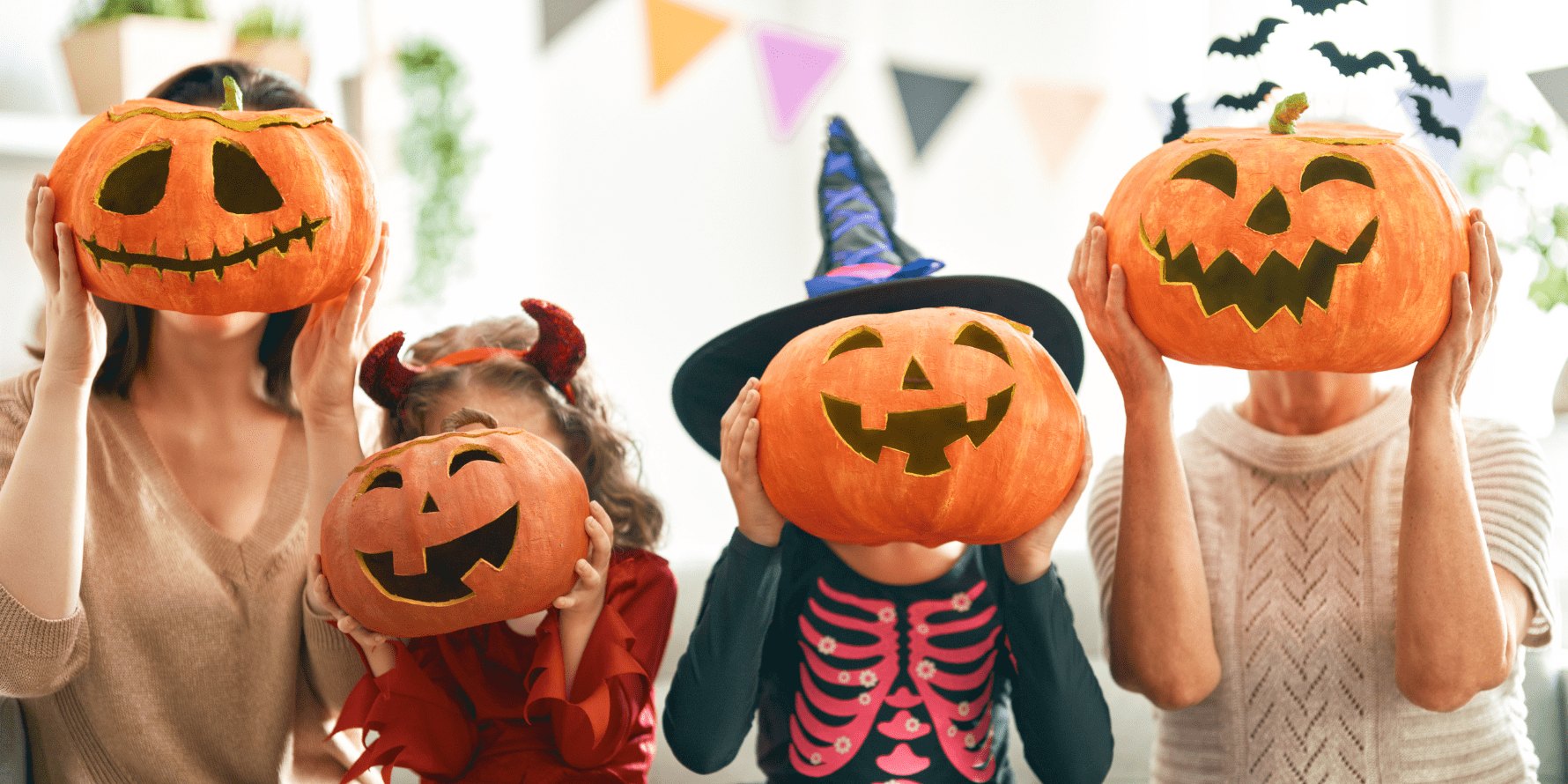 Two children in Halloween costumes hold carved jack-o'-lanterns in a decorated indoor space.