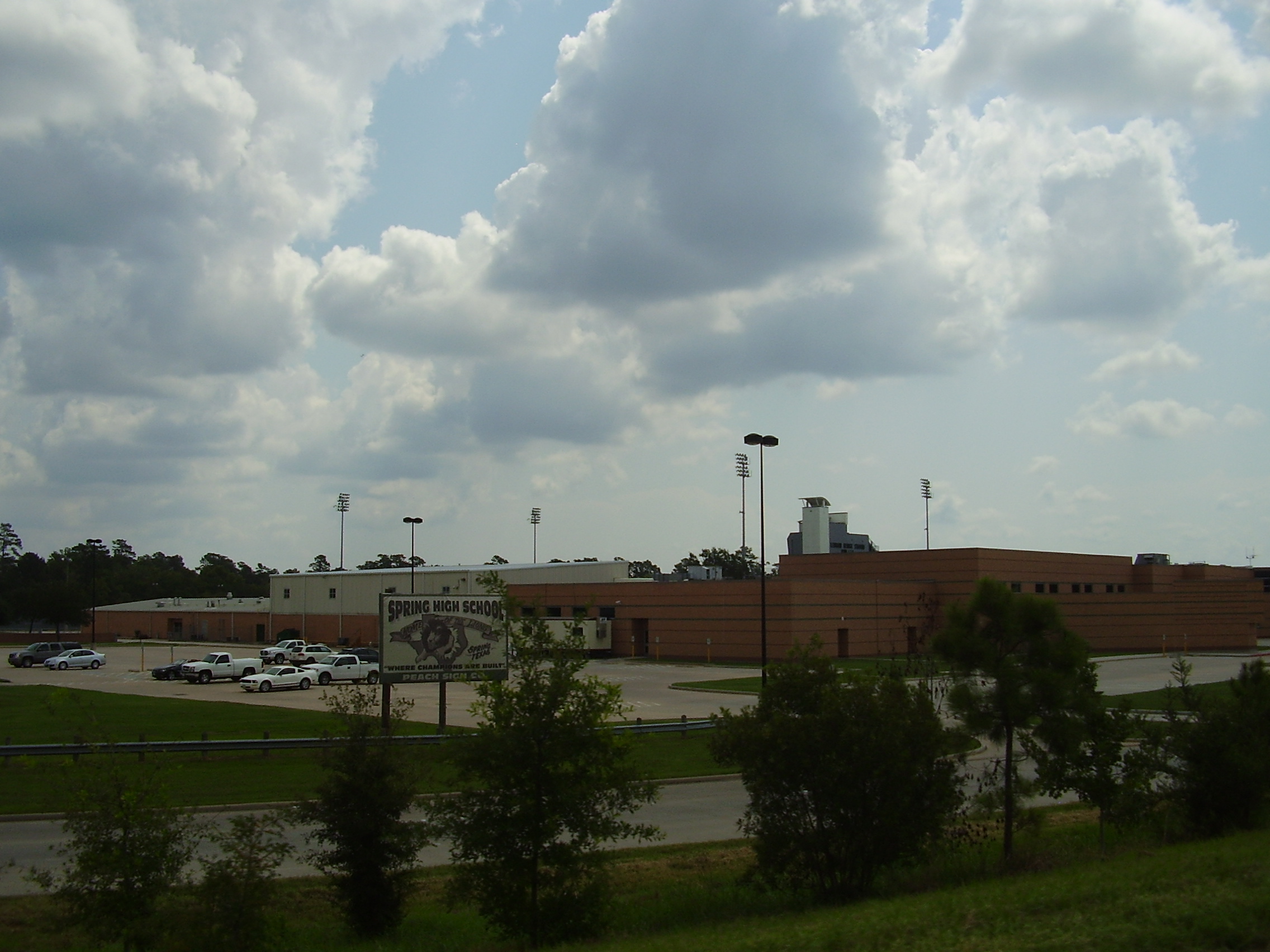 Brick high school building exterior with parking lot and sports field light towers visible.