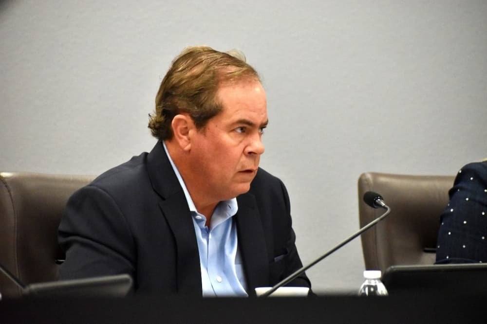 Man in dark blazer seated at a government meeting dais with a microphone.