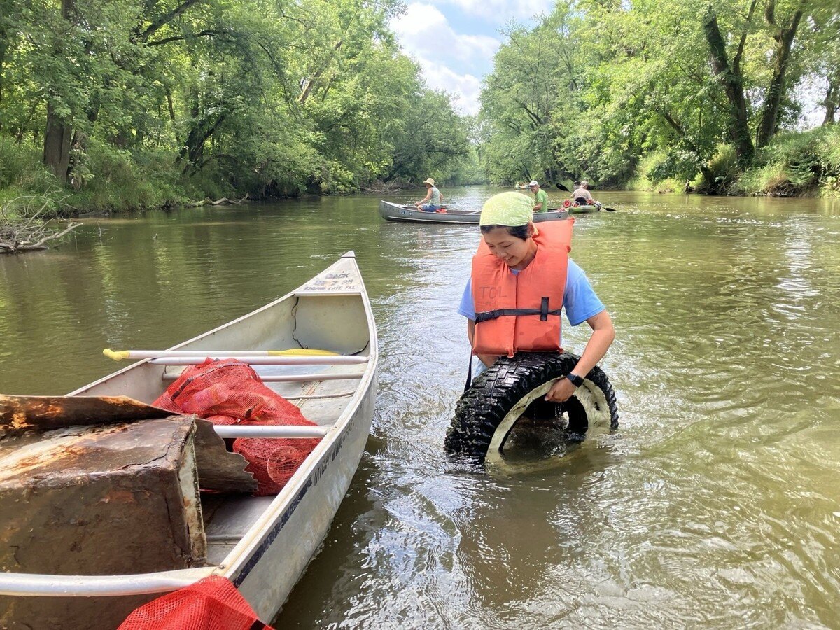 A volunteer in an orange life vest pulls a tire from a shallow river alongside a debris-filled canoe.