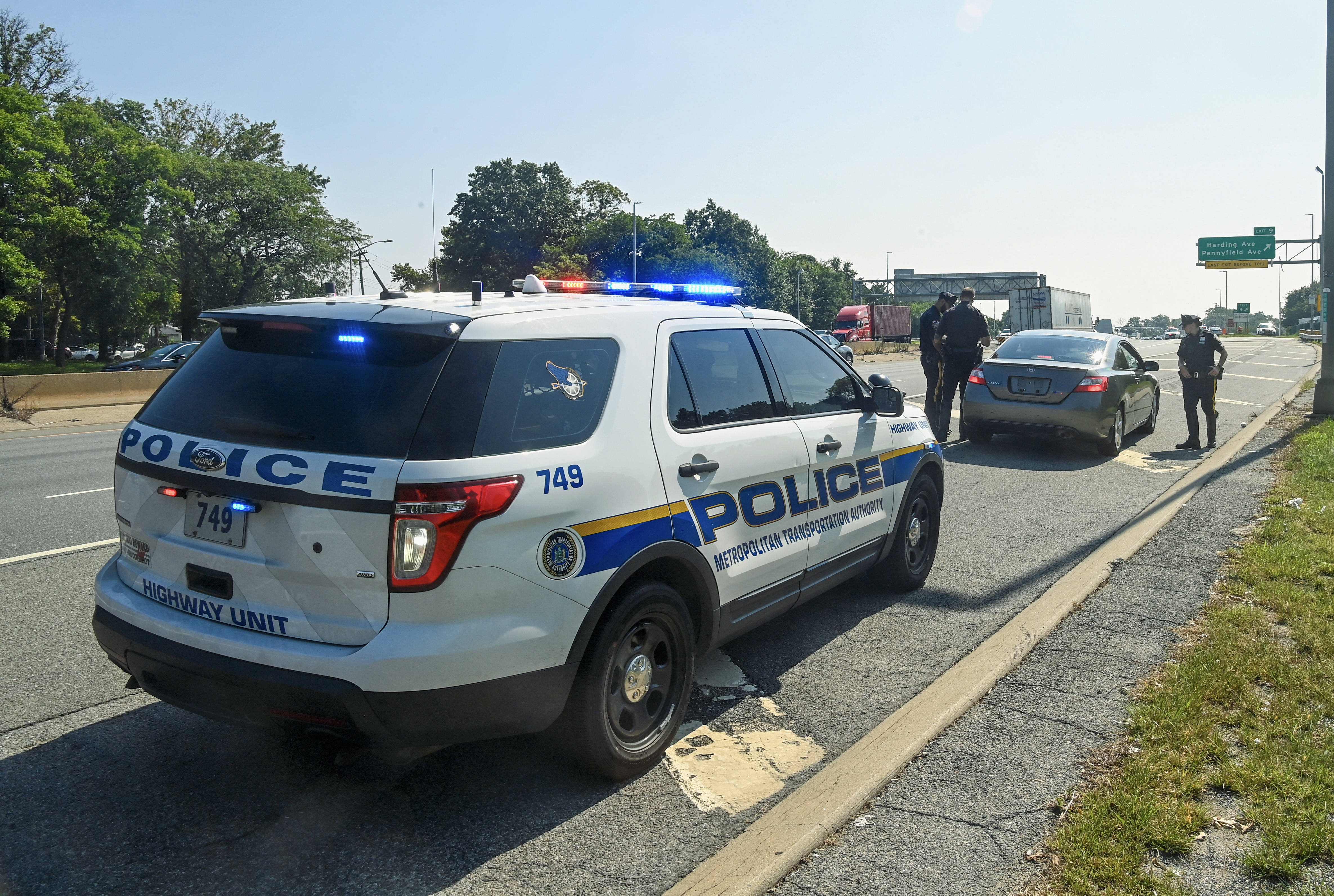 Police SUV with blue lights on, two officers standing near a stopped vehicle on a road shoulder.