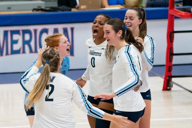 Five volleyball players in white and blue uniforms celebrate together on a court.