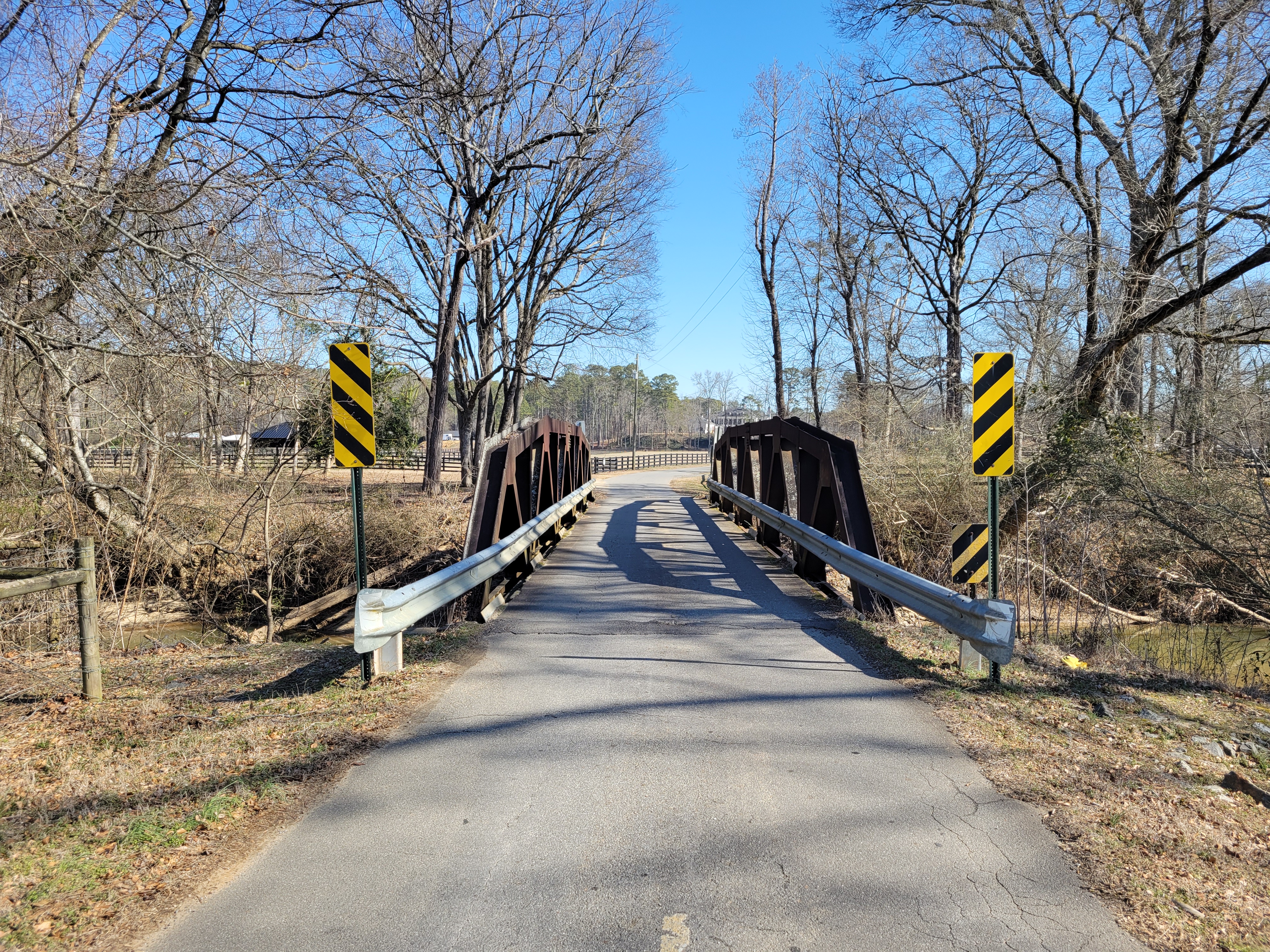 A narrow two-lane road bridge with metal truss railings crosses a wooded creek.