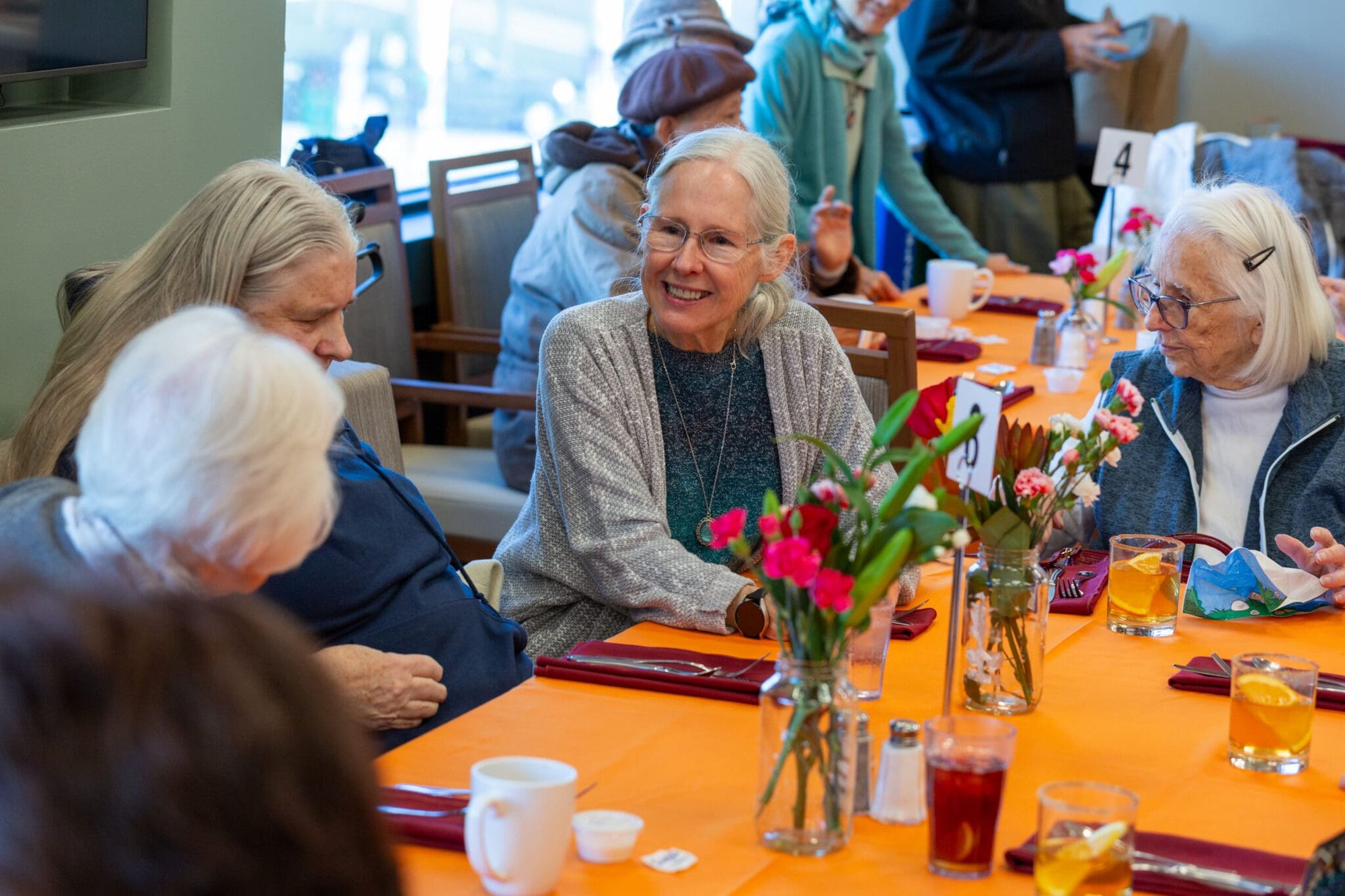 Older women seated around a flower-decorated table, talking and sharing drinks indoors.