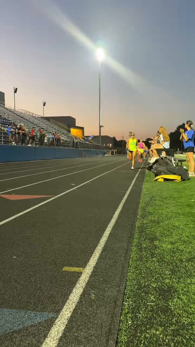 Female athletes stand near a lit running track at dusk with bleachers in the background.