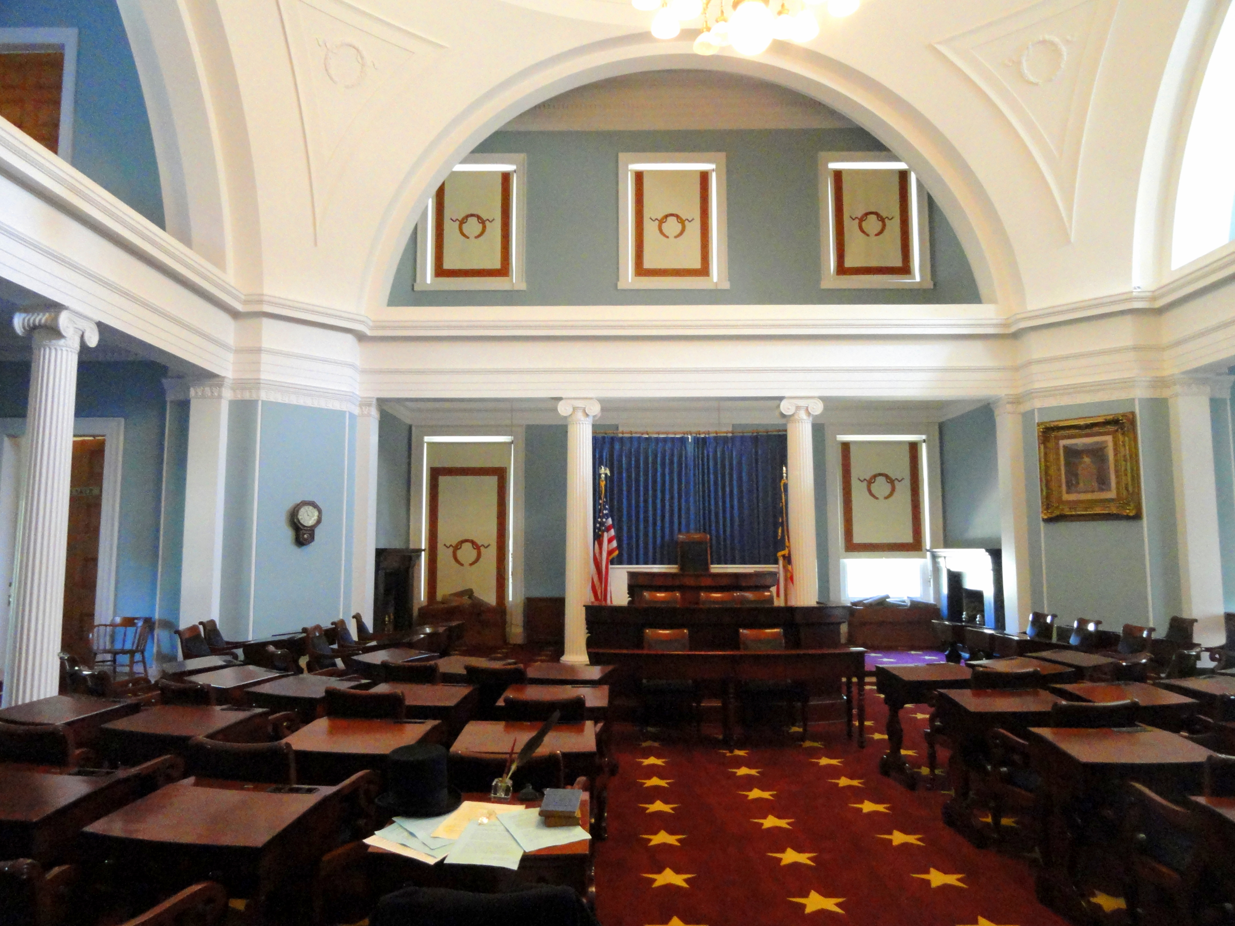 Rows of wooden desks face a raised dais inside a legislative chamber with white columns.