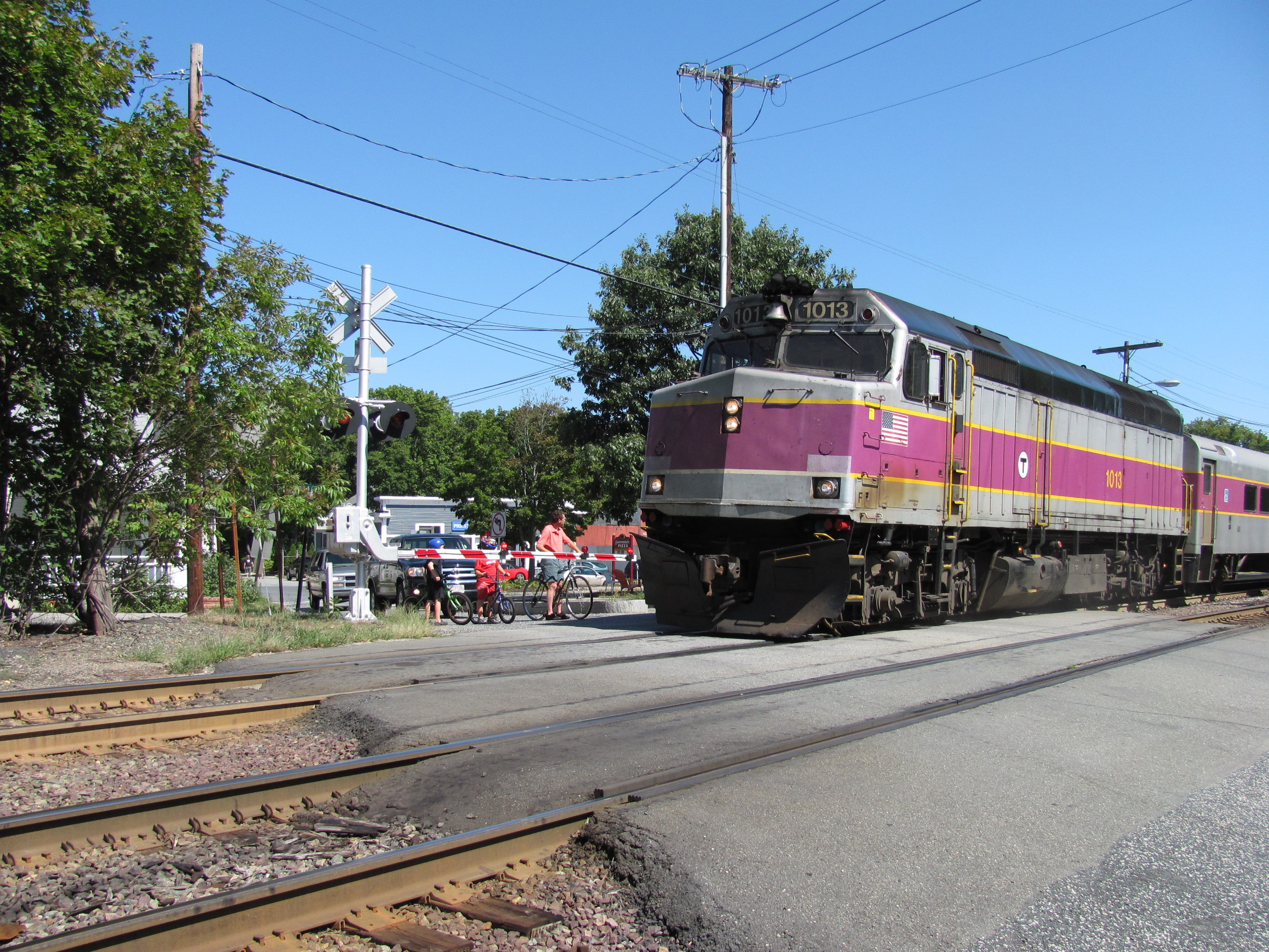Purple and silver MBTA Commuter Rail locomotive passing a grade crossing with cyclists waiting.