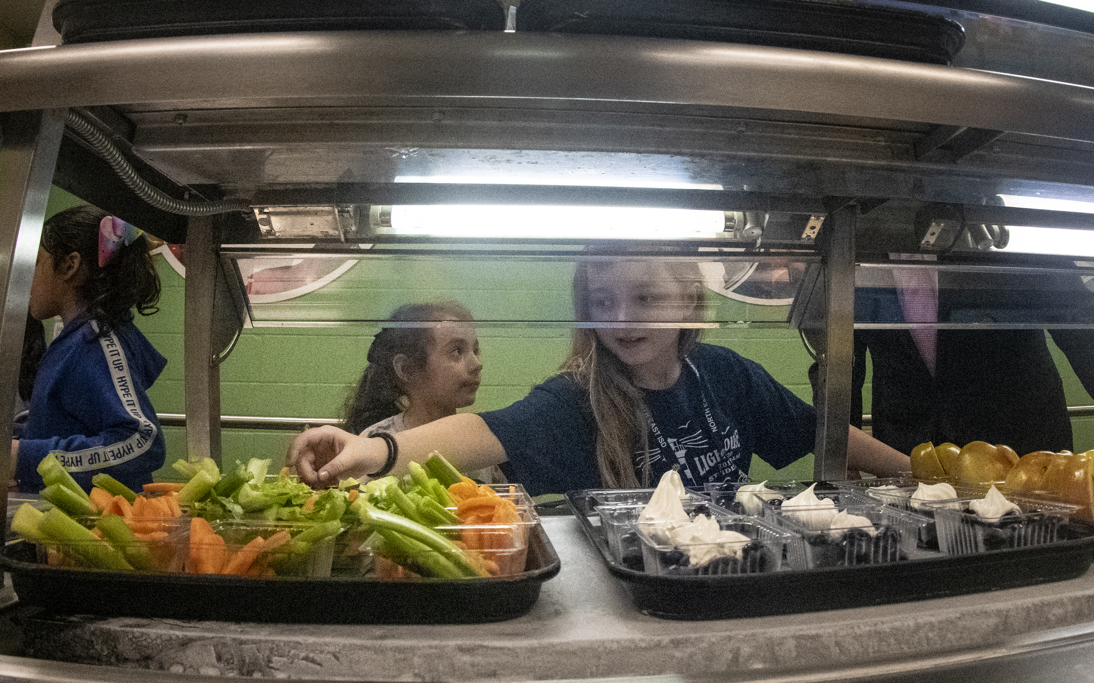 Two young girls selecting food at a school cafeteria serving line.
