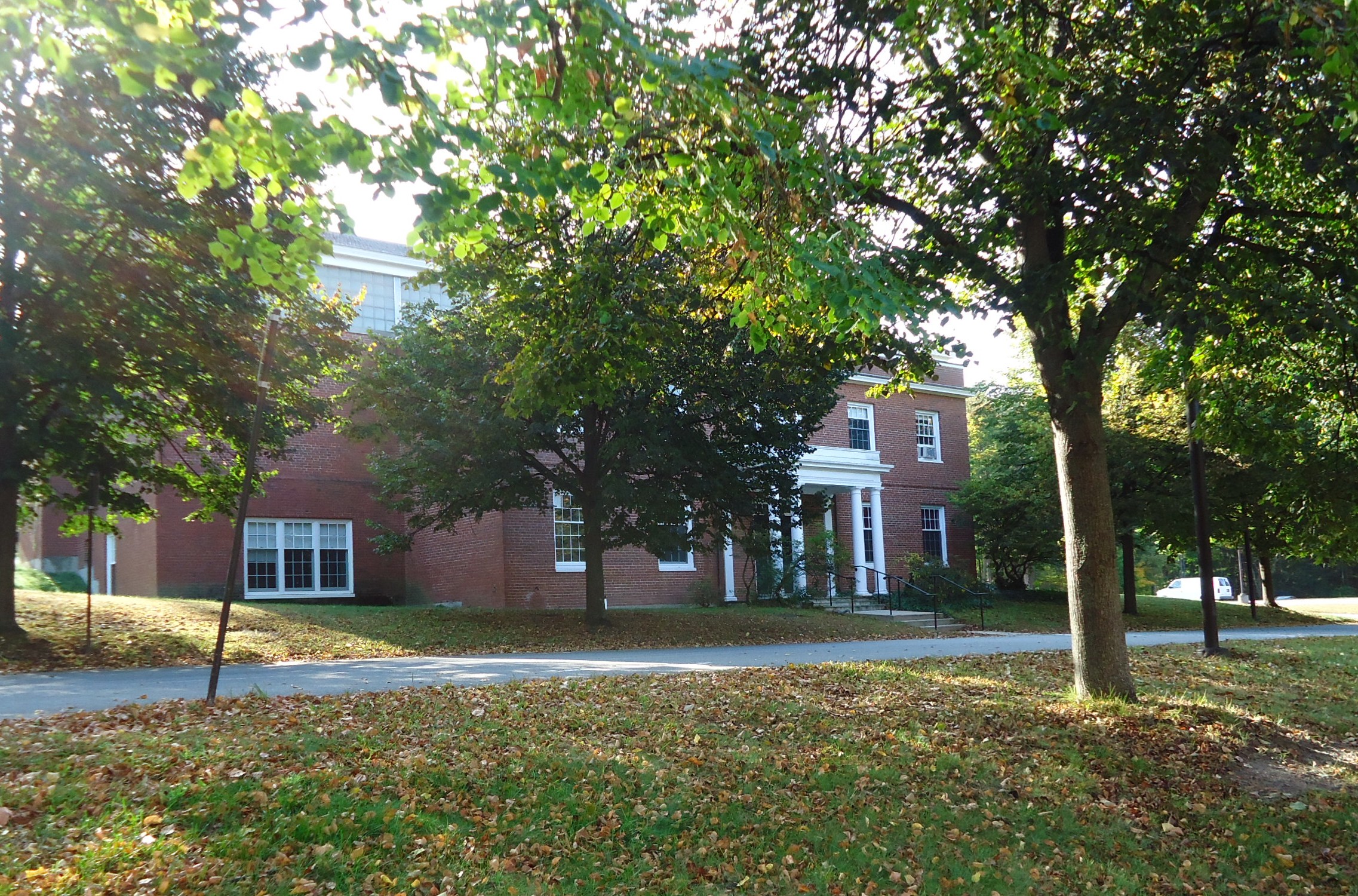 Brick campus building with white columned entrance seen through autumn trees, fallen leaves on lawn.