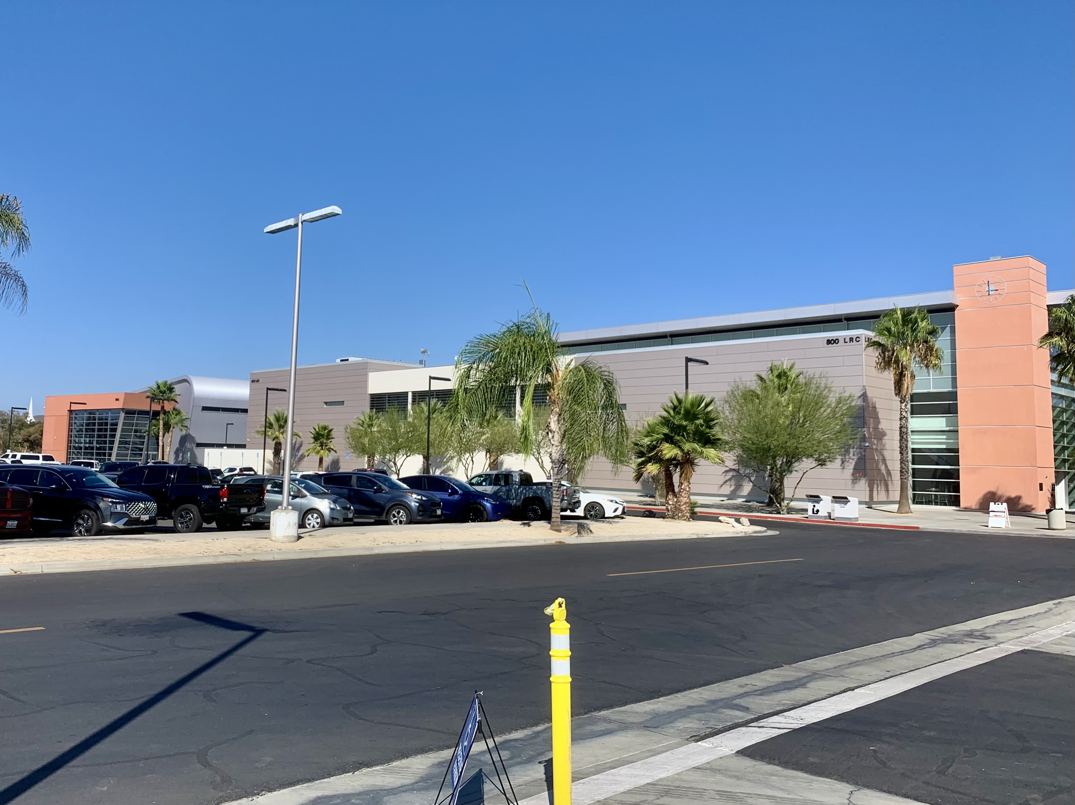 Modern college building with gray metal cladding, palm trees, and a sunny parking lot.