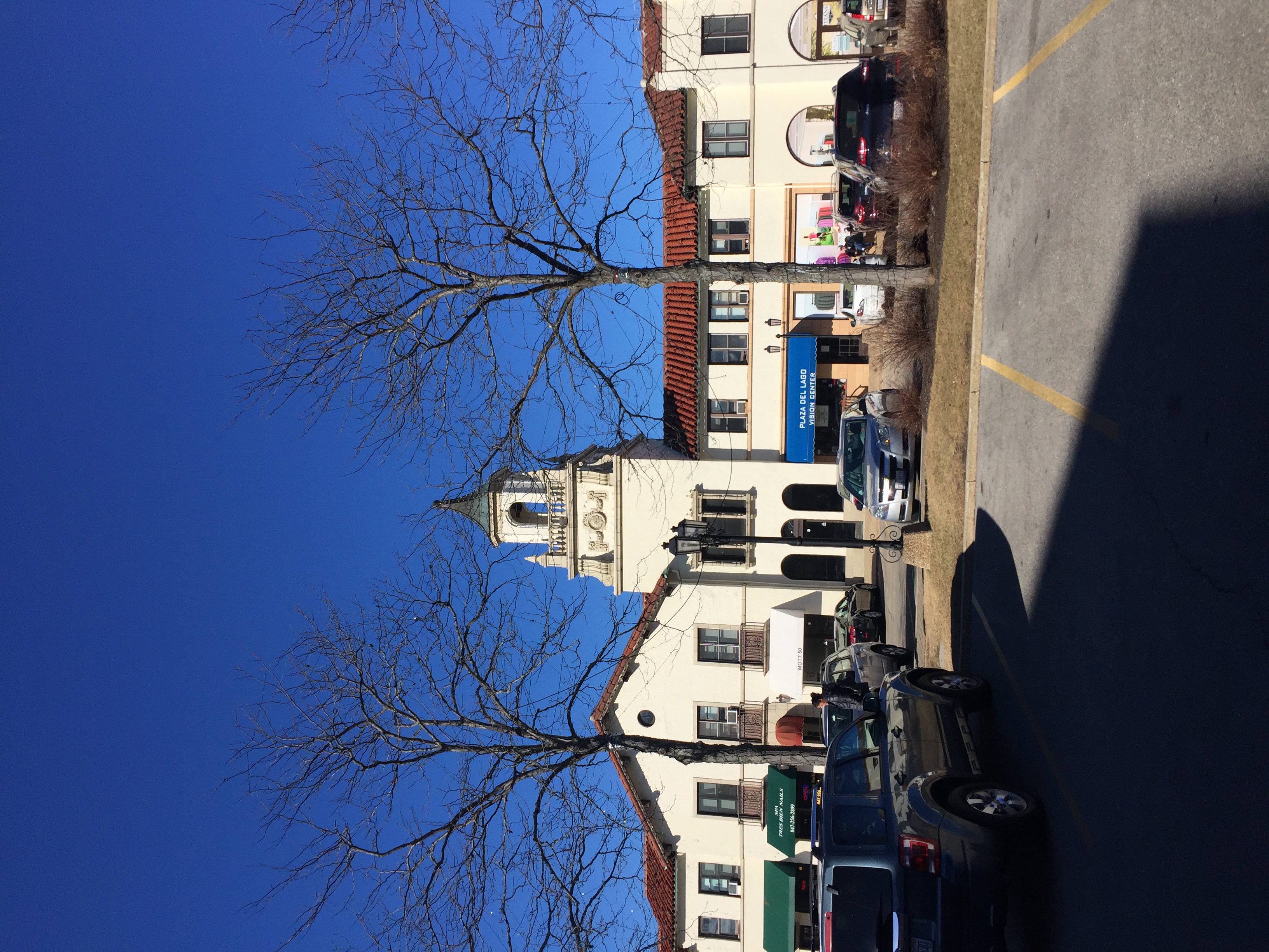 White Spanish Revival shopping center with bell tower and blue awning, parking lot in foreground.