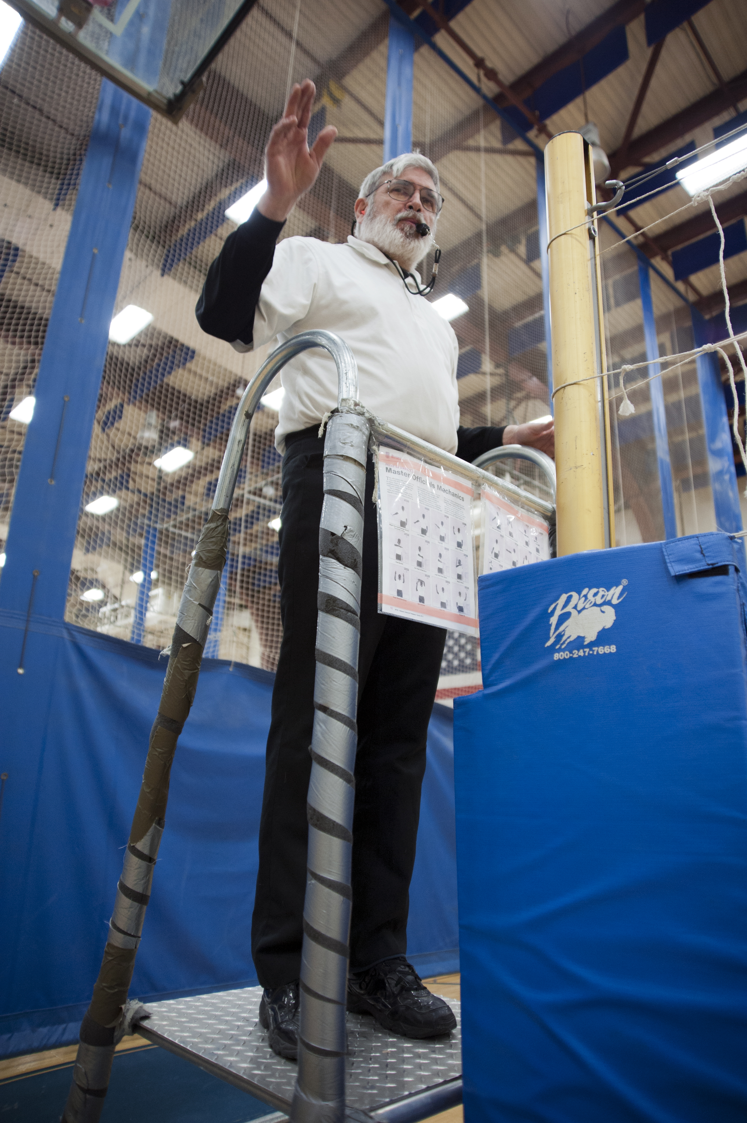 A referee on an elevated stand raises one hand to signal during an indoor volleyball game.