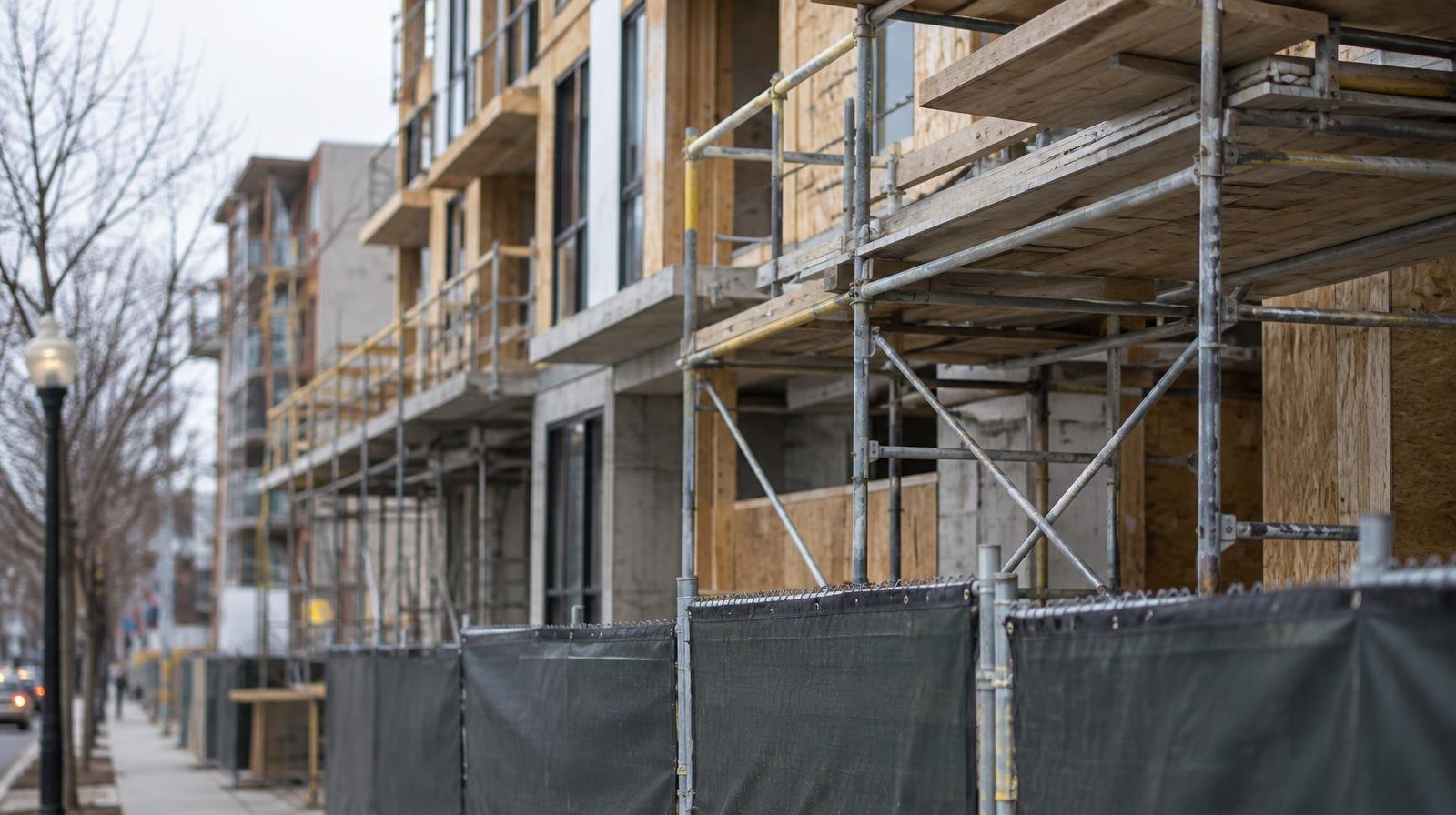 Multi-story apartment building under construction with scaffolding along a city sidewalk.