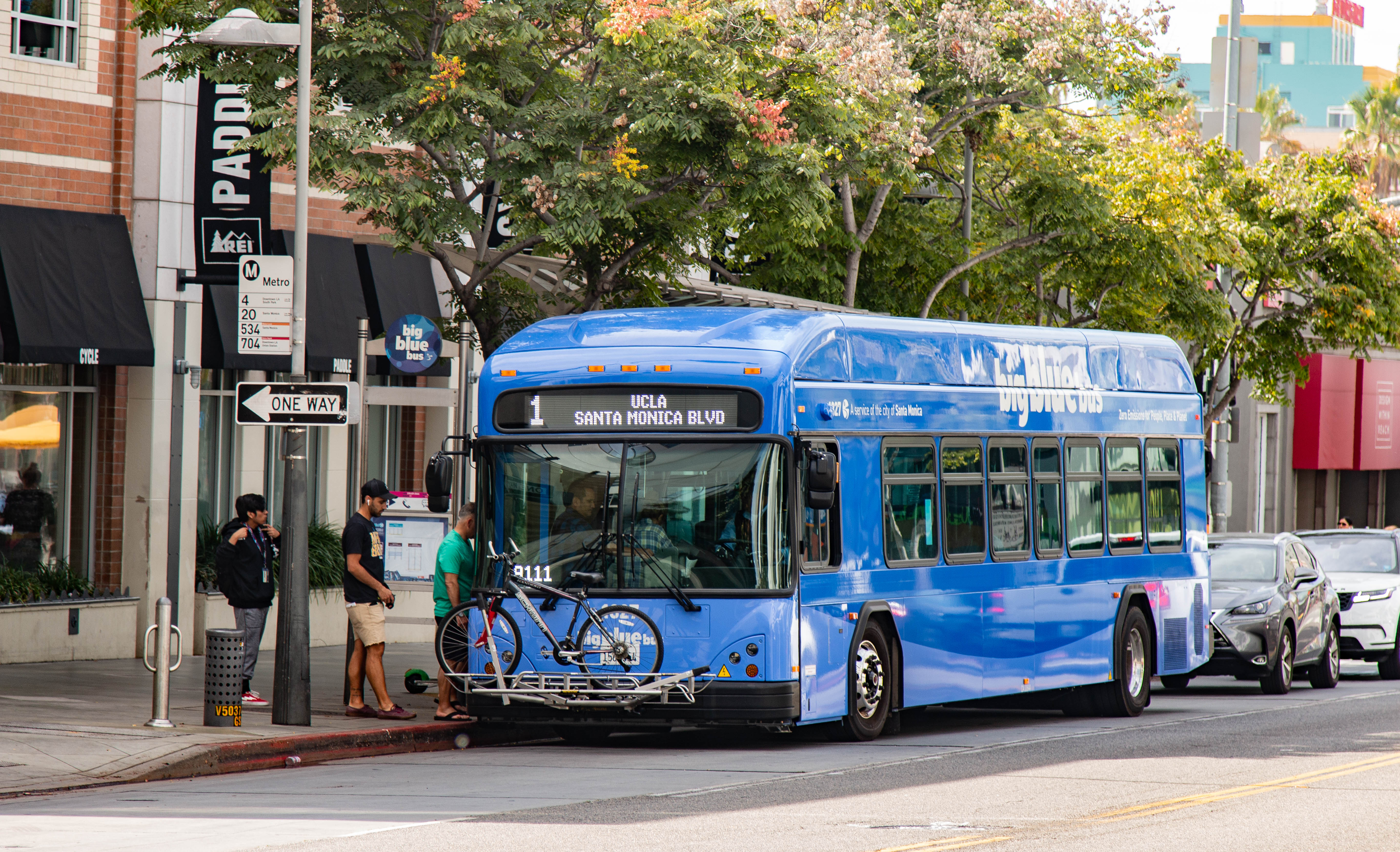Blue city bus stopped at a street corner with passengers boarding and a bicycle on the front rack.