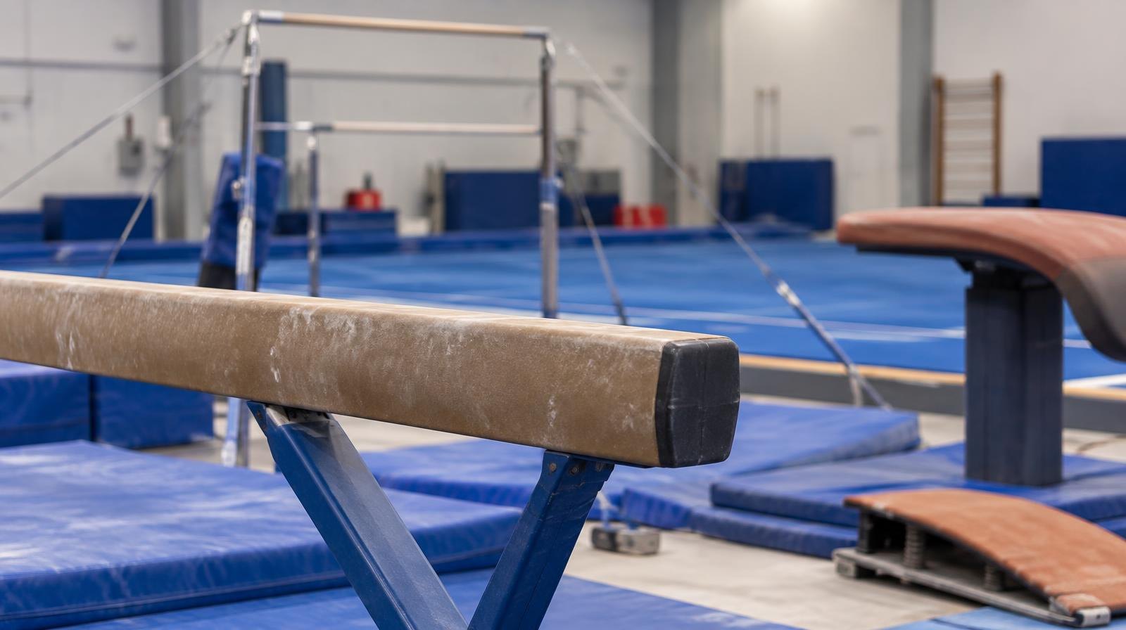 A balance beam, uneven bars, and vault table arranged in a bright gymnasium.