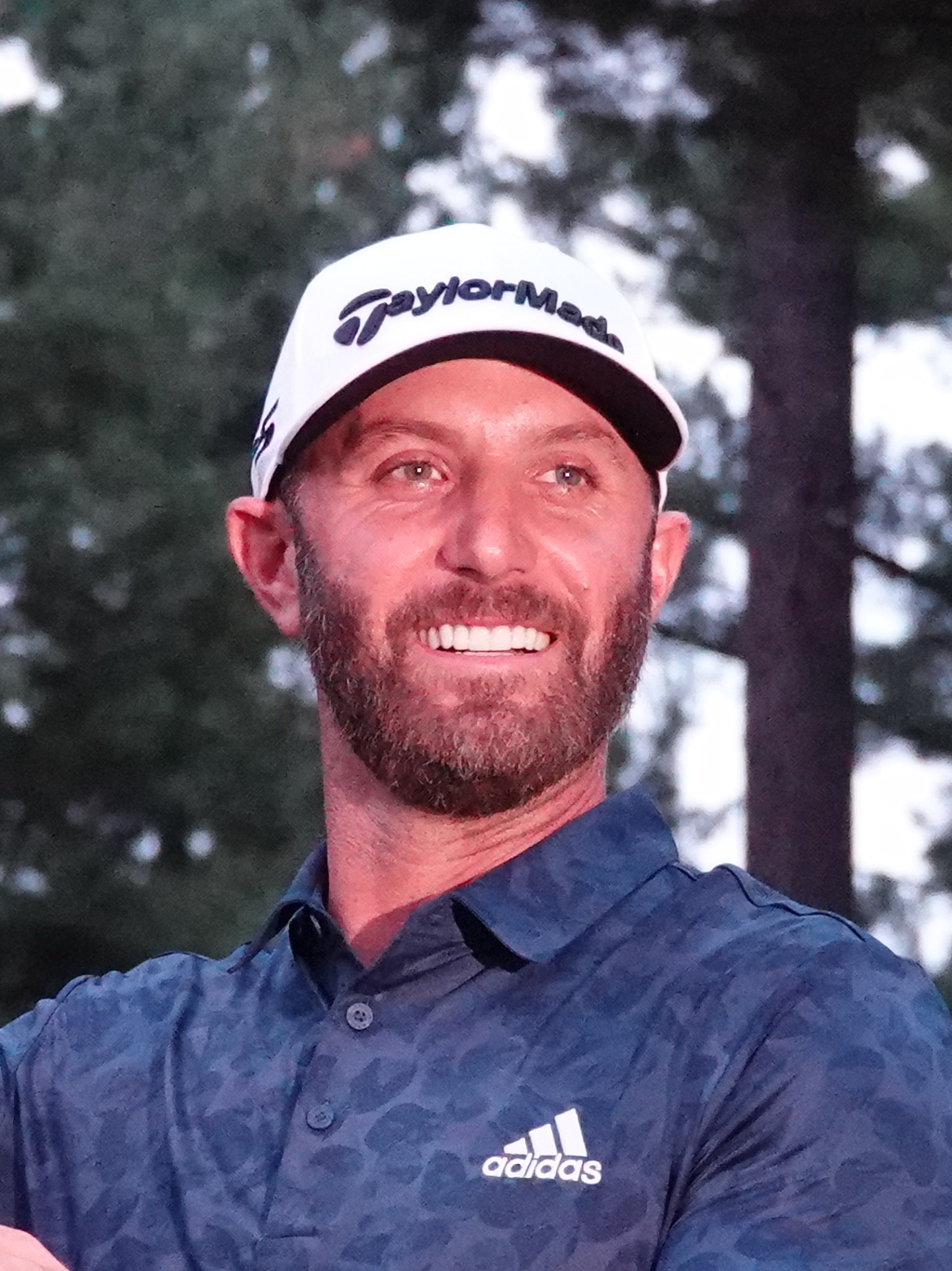 Smiling man in a white golf cap and blue polo shirt outdoors with trees behind him.