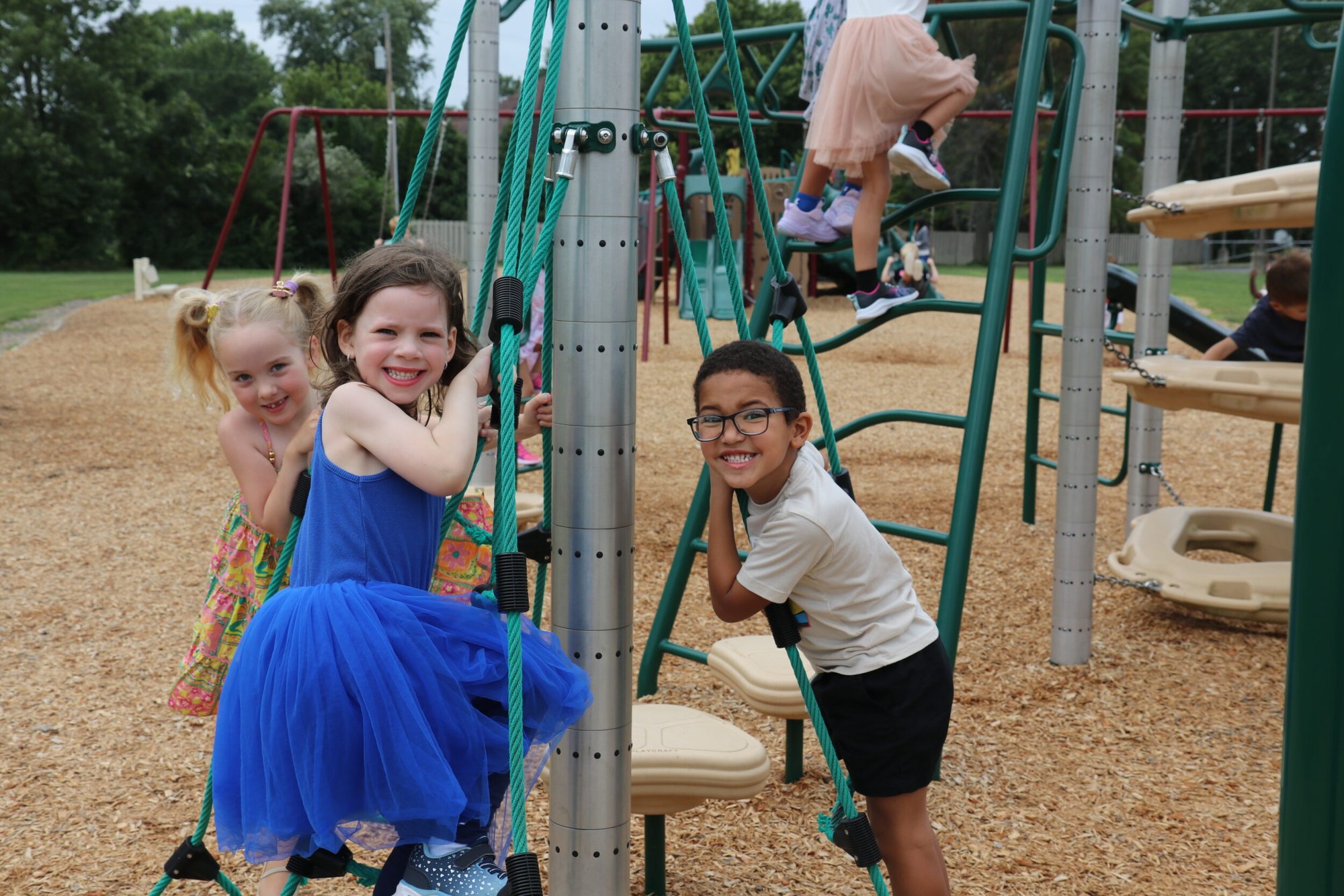 Three young girls smiling and climbing on a green playground structure.